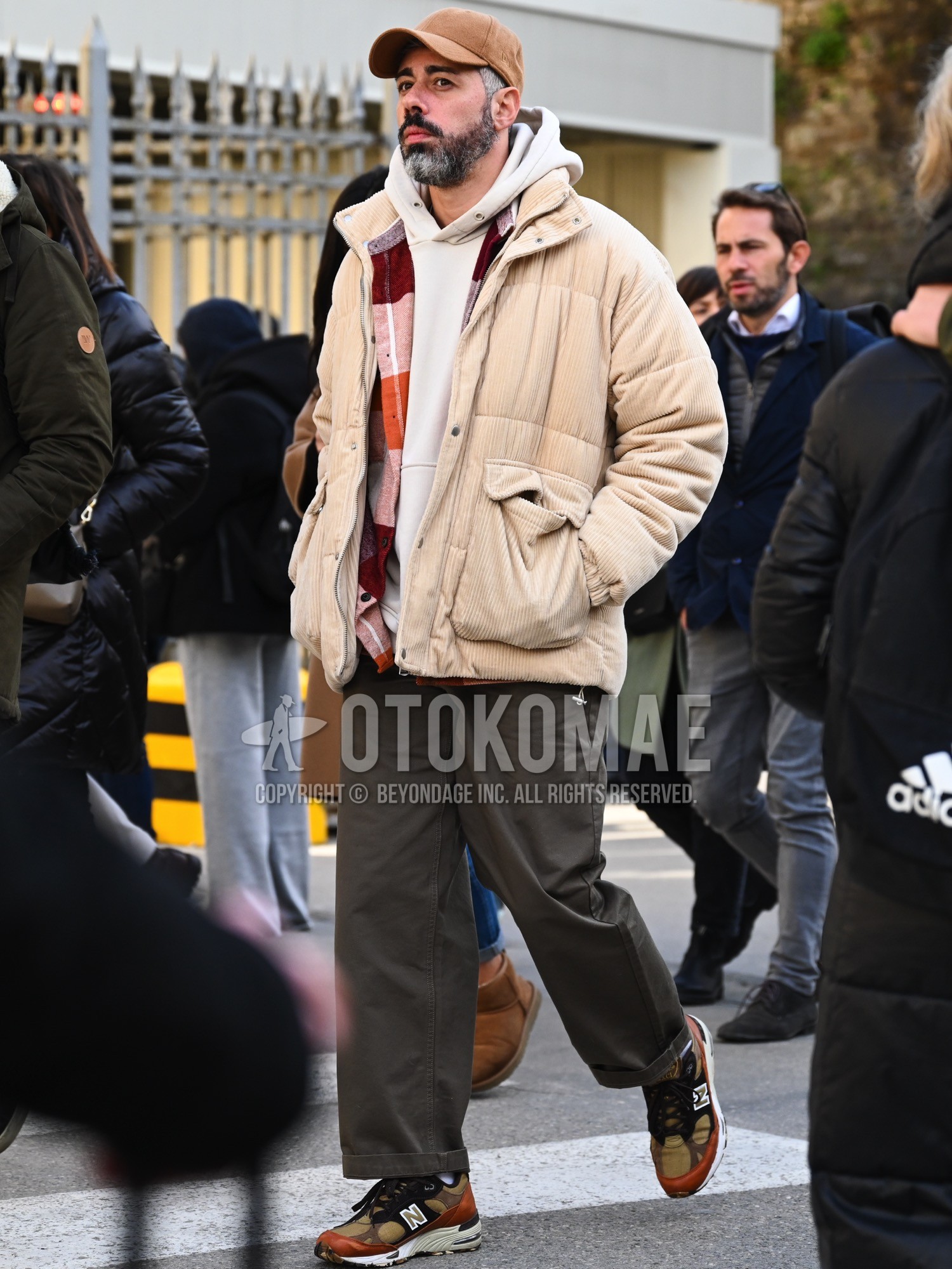 Men's autumn winter outfit with brown plain baseball cap, beige plain down jacket, white plain hoodie, multi-color check shirt, gray plain cotton pants, olive green low-cut sneakers.