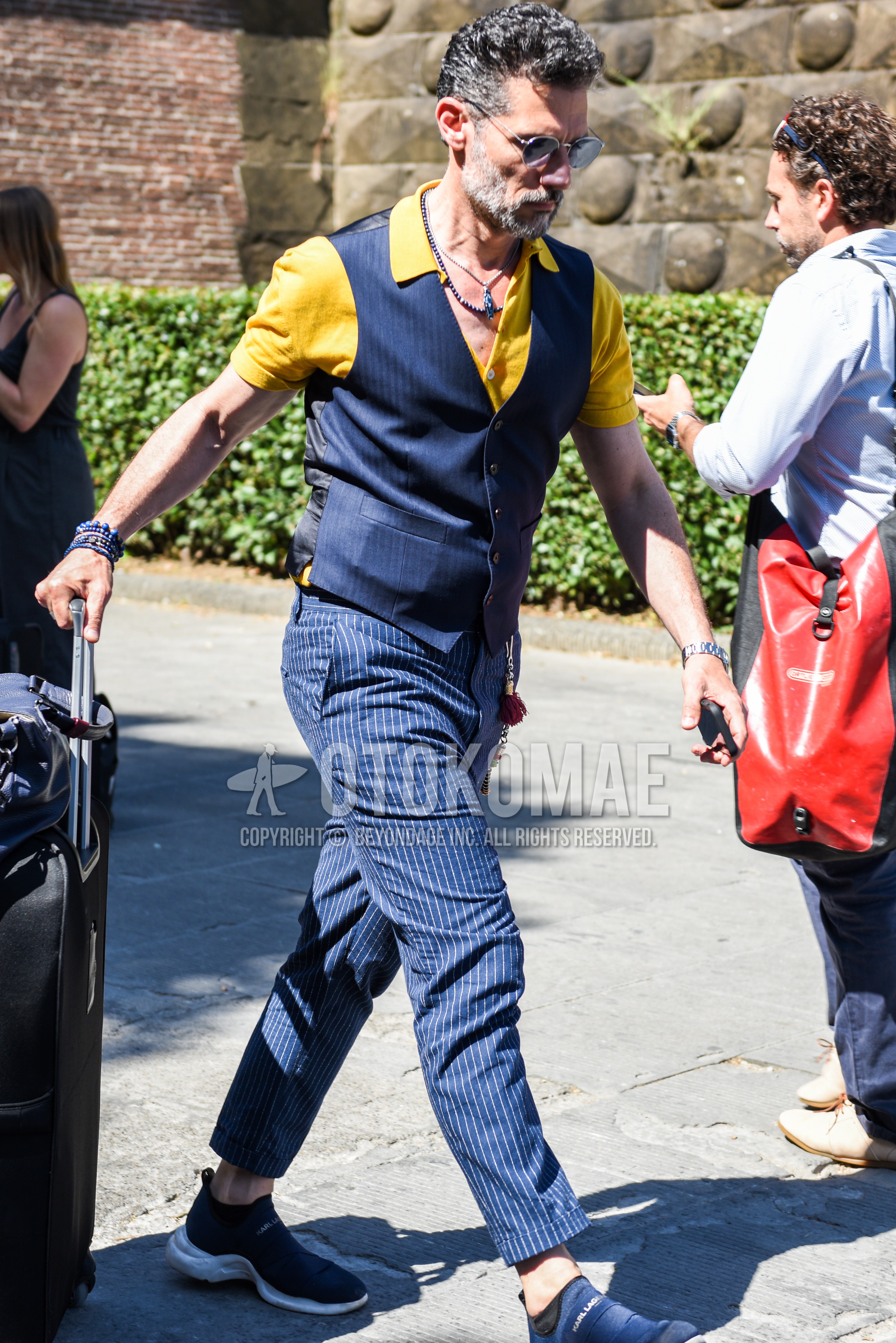 Men's spring summer outfit with silver plain sunglasses, yellow plain polo shirt, navy stripes gilet, blue plain slacks, navy low-cut sneakers.