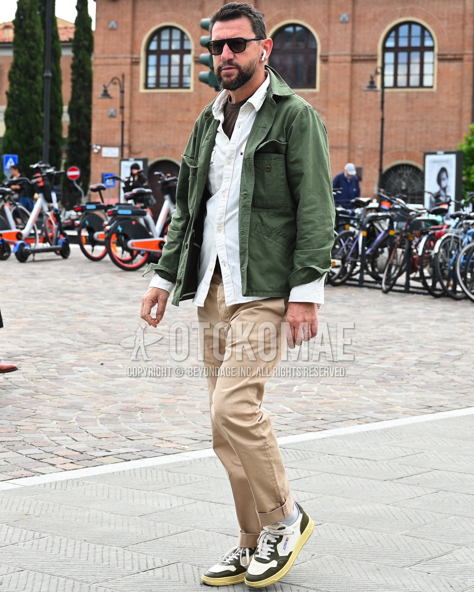 Men's spring summer autumn outfit with black plain sunglasses, olive green plain military jacket, white plain shirt, beige plain chinos, white low-cut sneakers.
