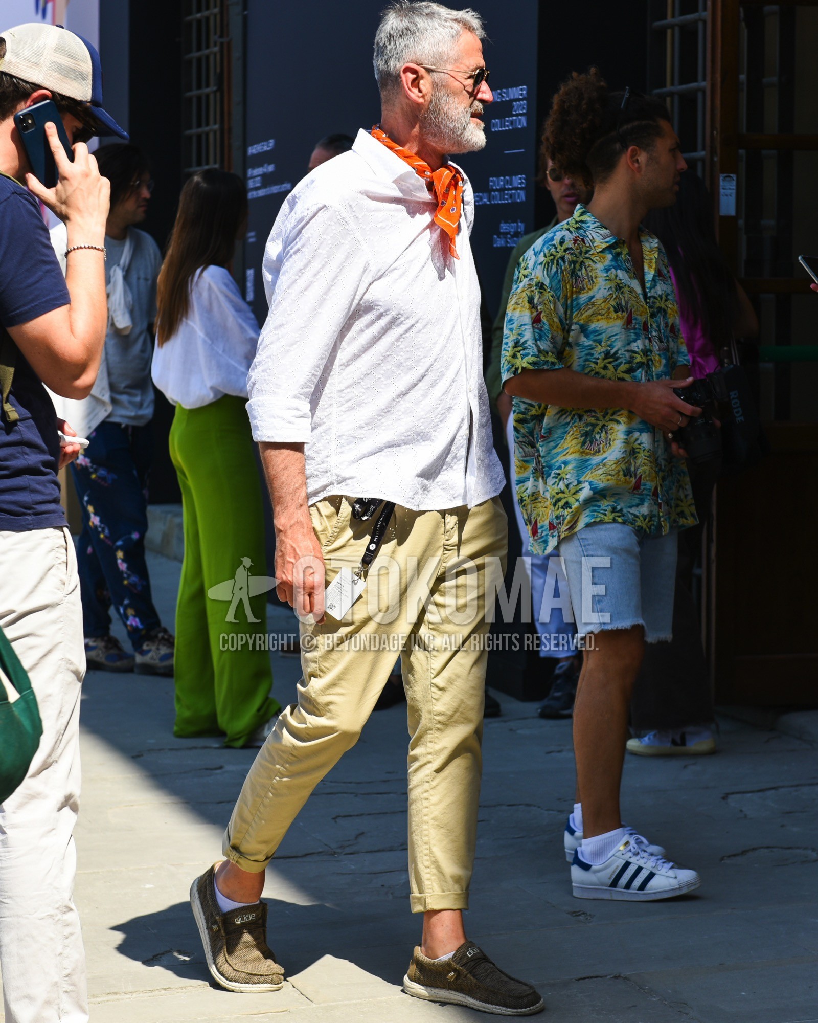 Men's spring summer outfit with gold plain sunglasses, orange scarf bandana/neckerchief, white plain shirt, beige plain chinos, white plain socks, brown slip-on sneakers.