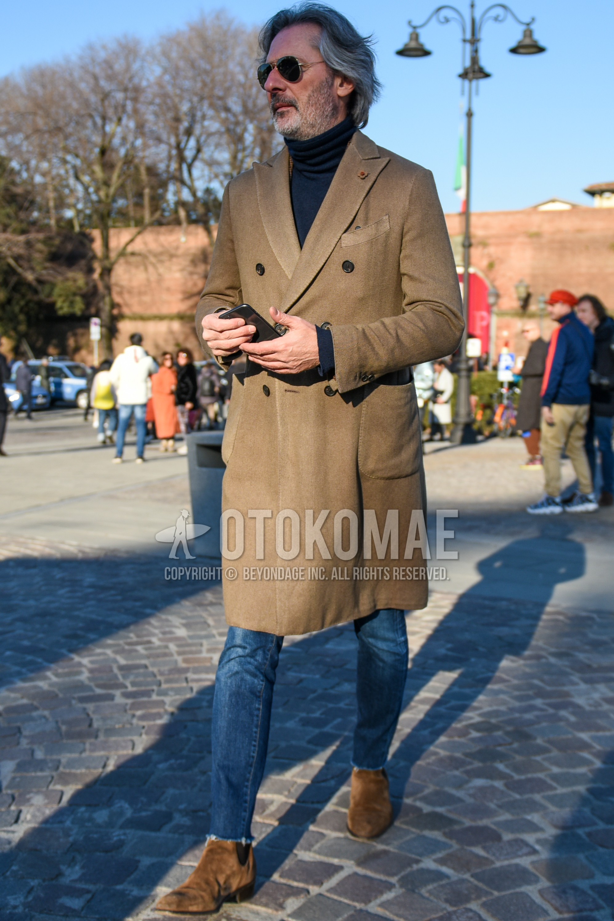 Men's autumn winter outfit with silver plain sunglasses, beige plain chester coat, black plain turtleneck knit, blue plain denim/jeans, blue plain cropped pants, brown side-gore boots.