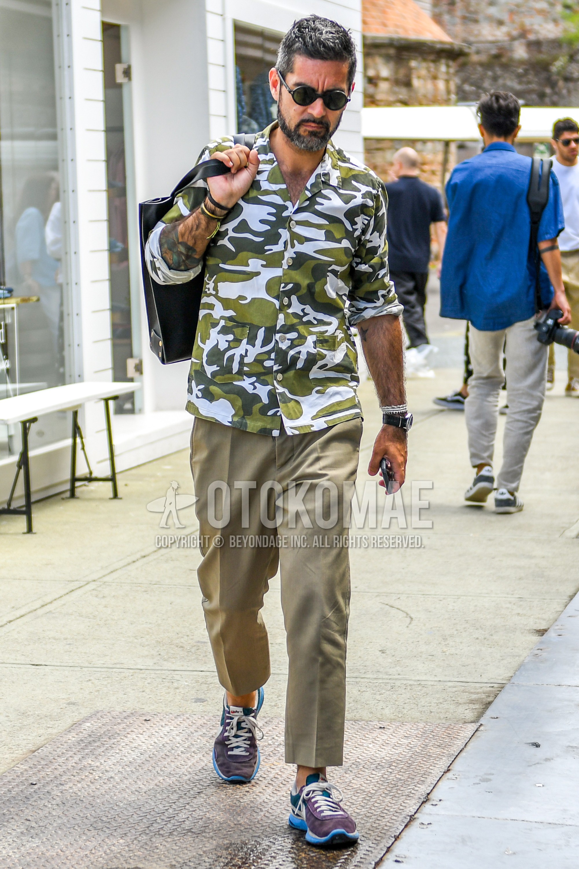 Men's spring summer outfit with plain sunglasses, olive green white camouflage shirt, beige plain chinos, beige plain ankle pants, beige plain slacks, multi-color low-cut sneakers, black plain briefcase/handbag.