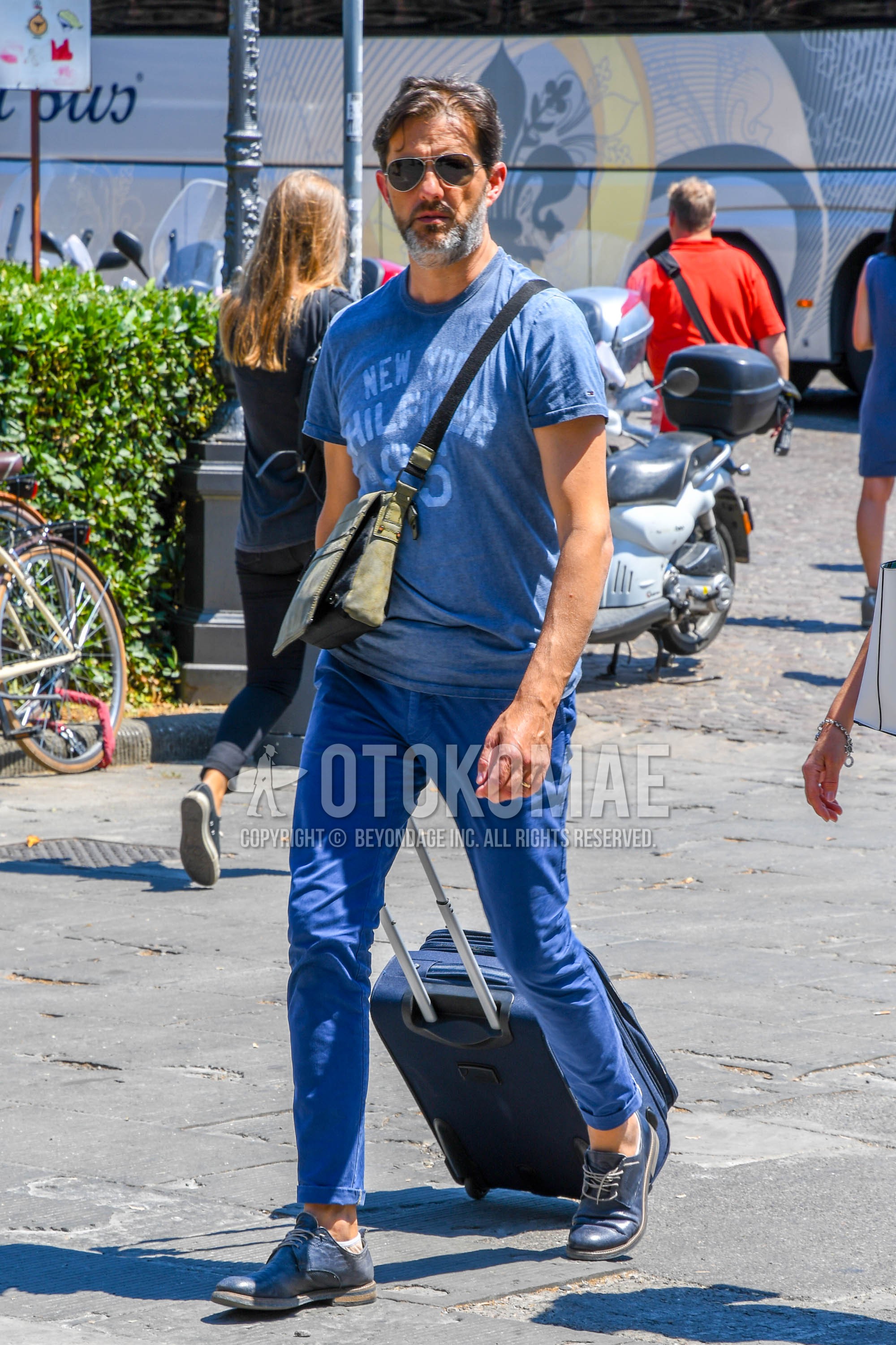 Men's summer outfit with plain sunglasses, navy graphic t-shirt, blue plain chinos, blue chukka boots, olive green plain shoulder bag.