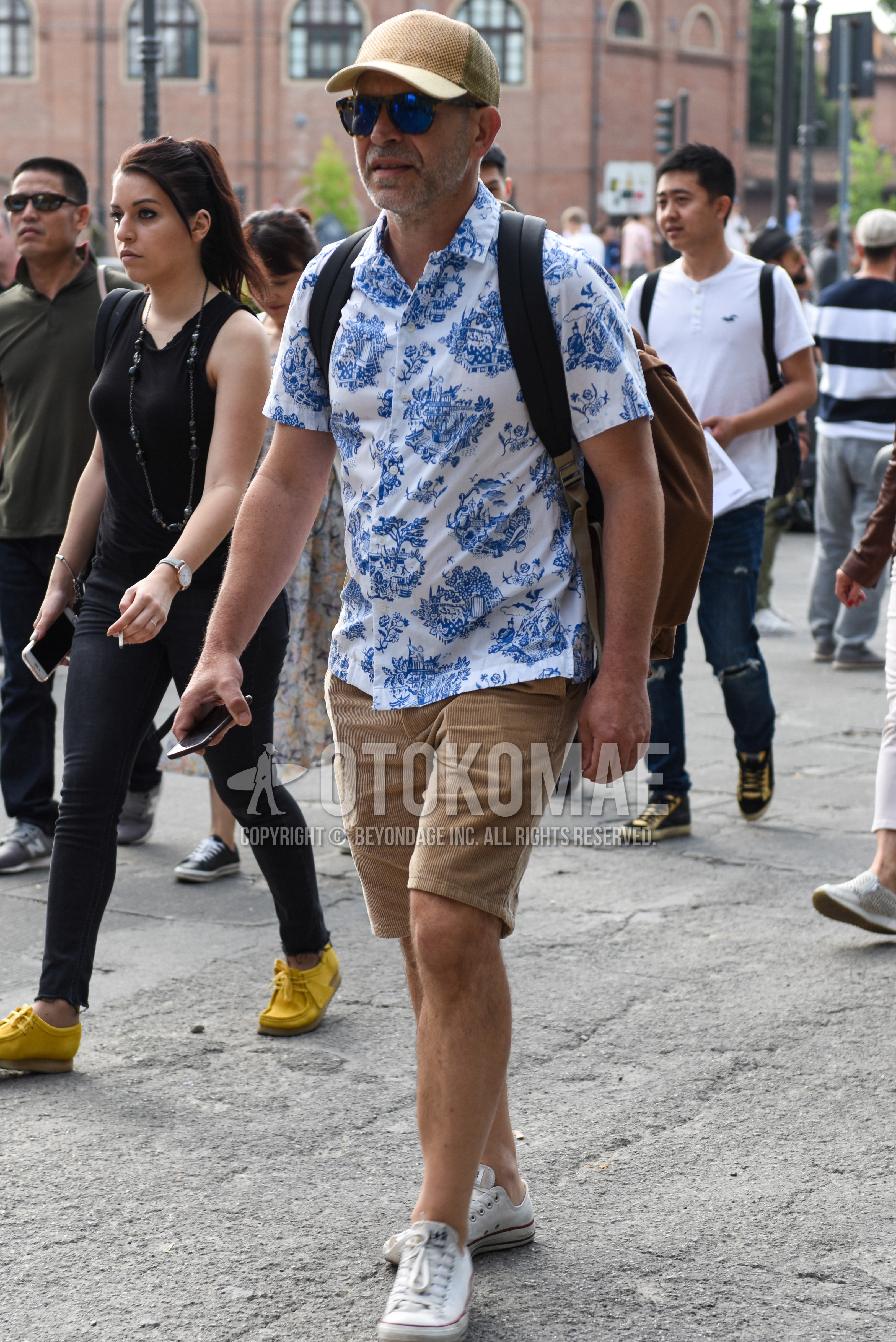 Men's summer outfit with beige plain baseball cap, black tortoiseshell sunglasses, white graphic shirt, beige plain short pants, white low-cut sneakers.
