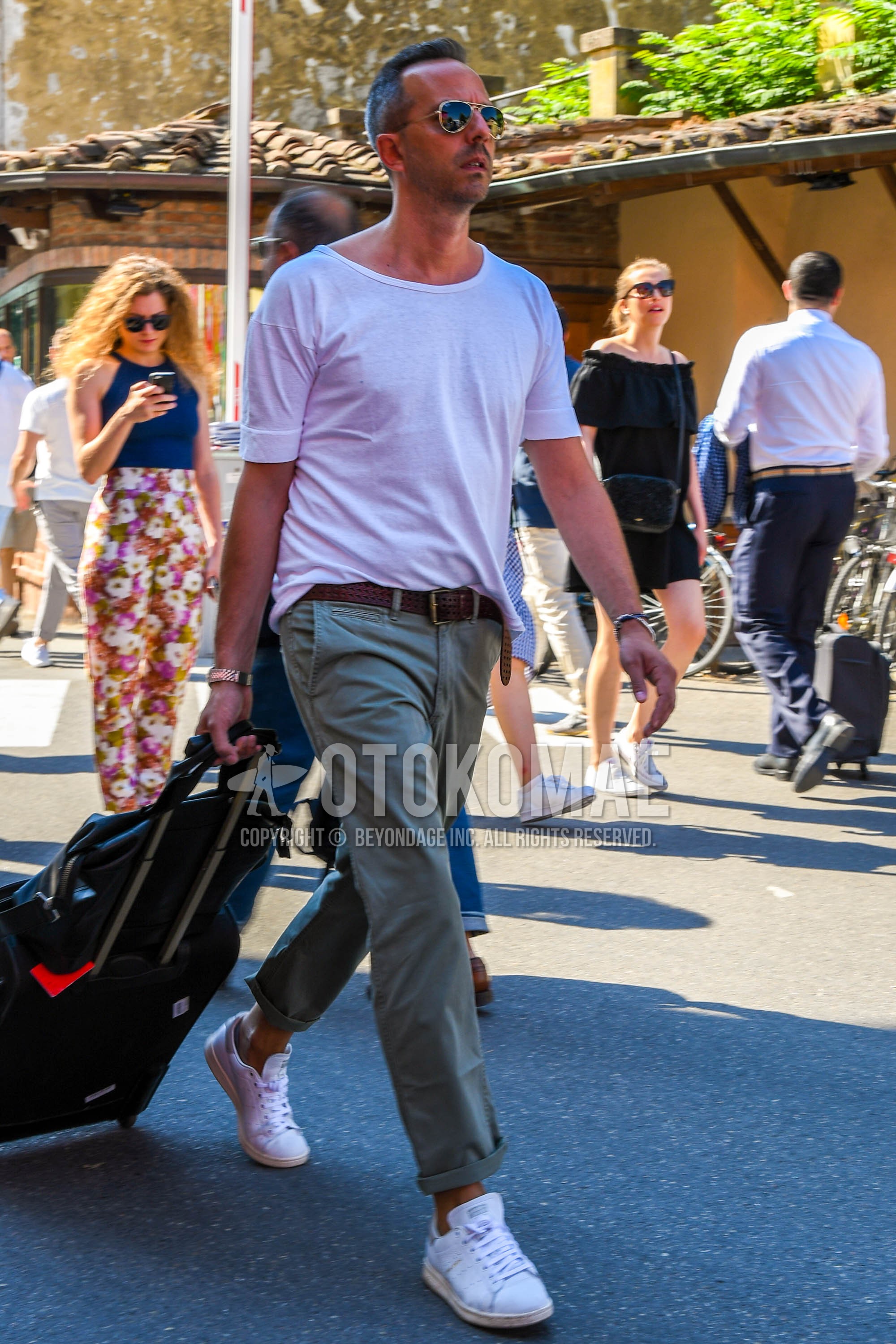 Men's spring summer outfit with gold plain sunglasses, white plain t-shirt, brown plain leather belt, olive green plain chinos, white low-cut sneakers, black plain suitcase.