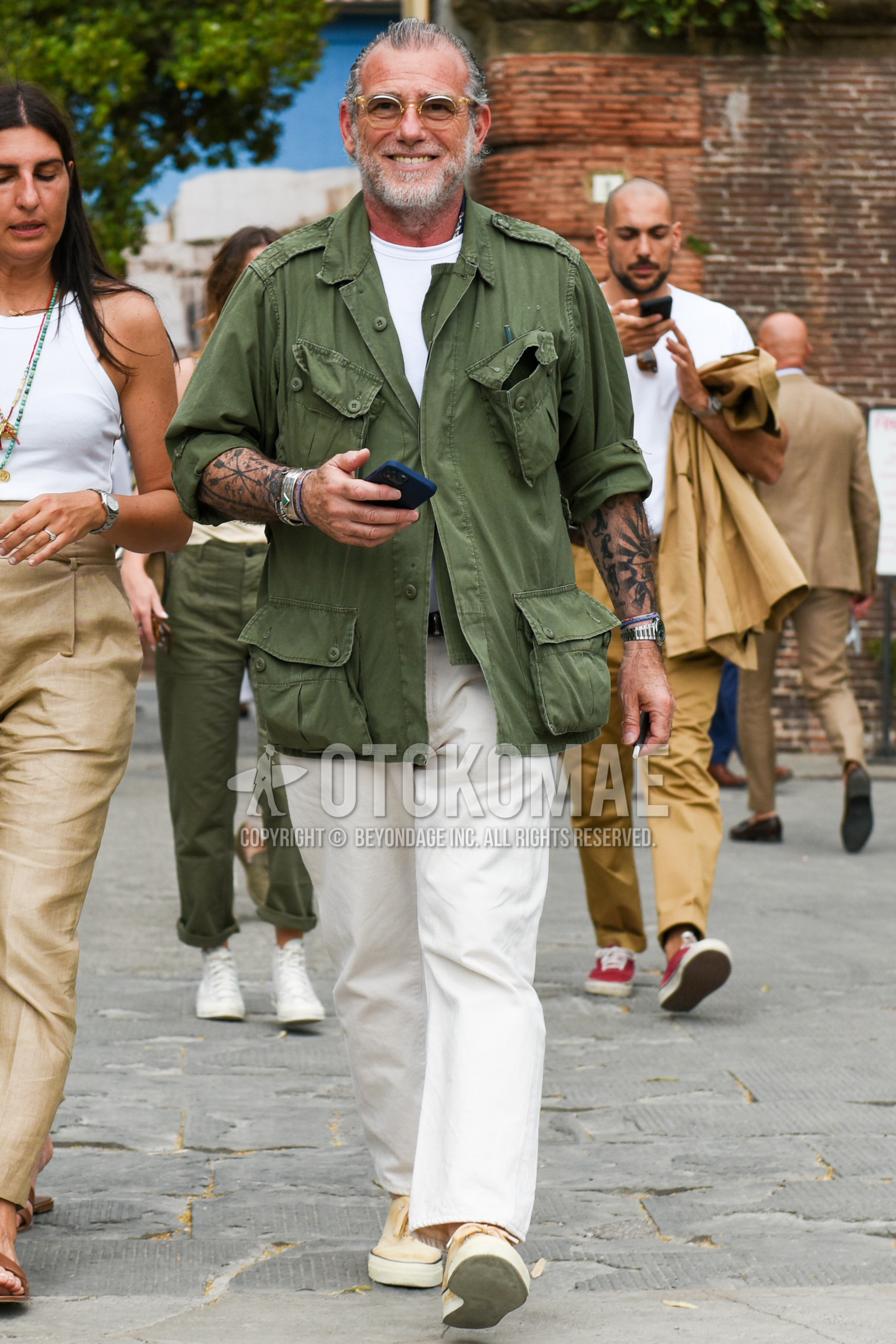 Men's spring summer outfit with clear plain glasses, olive green plain field jacket/hunting jacket, white plain t-shirt, white plain cotton pants, yellow low-cut sneakers.