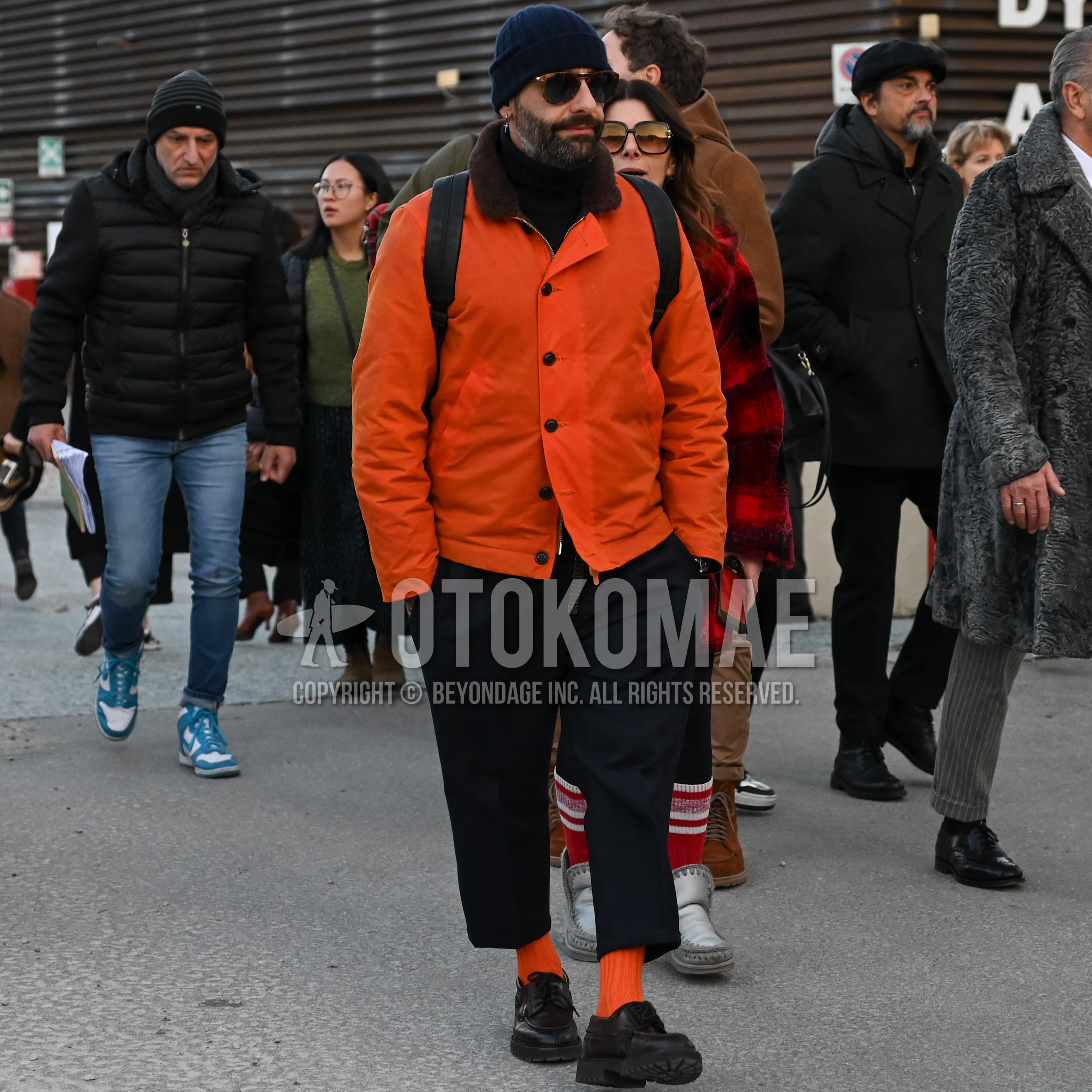 Men's autumn winter outfit with navy plain knit cap, tortoiseshell sunglasses, orange plain coach jacket, black plain ankle pants, orange plain socks, black u-tip shoes leather shoes, black plain backpack.