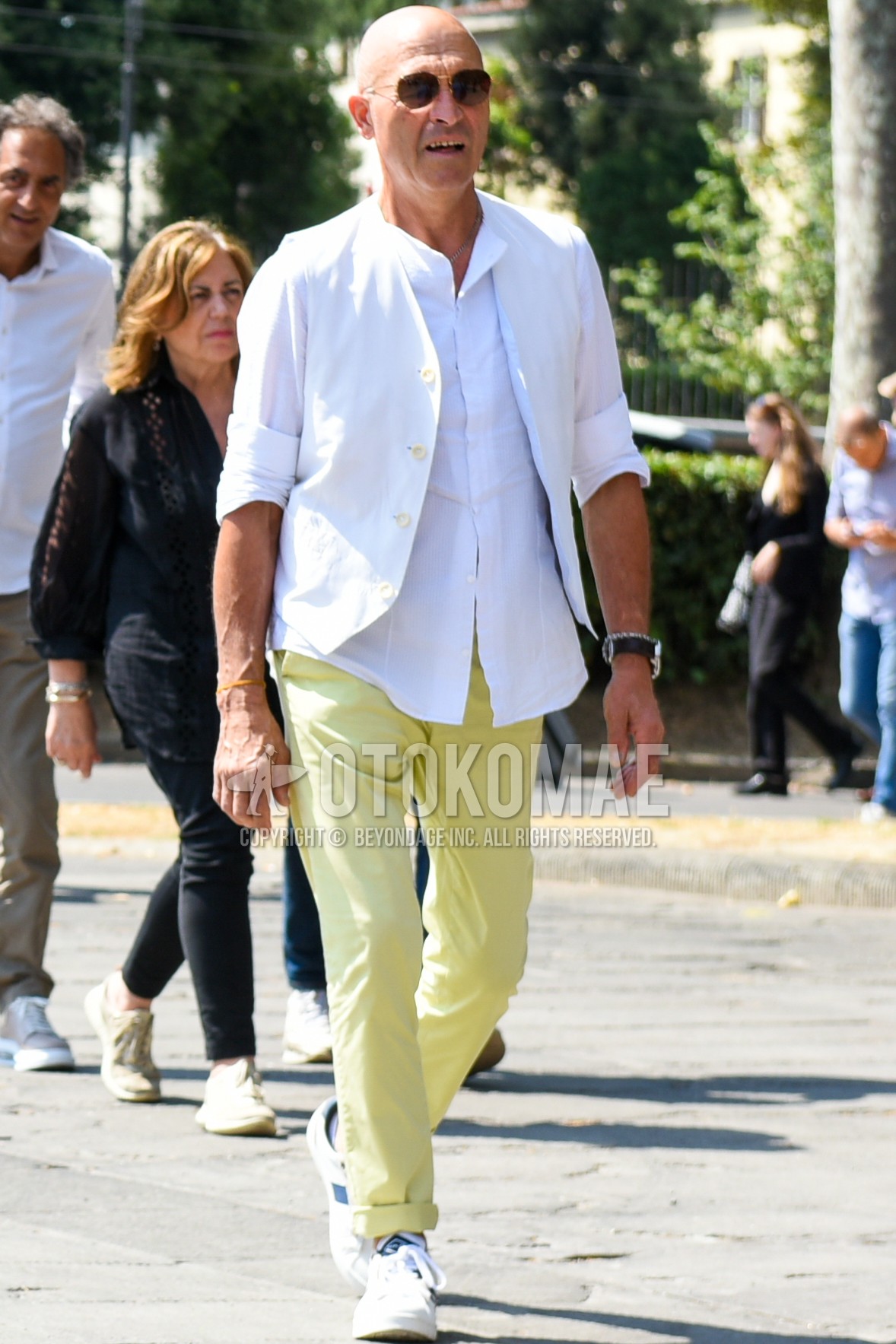 Men's spring summer outfit with gold plain sunglasses, white plain casual vest, white plain shirt, yellow plain chinos, white low-cut sneakers.