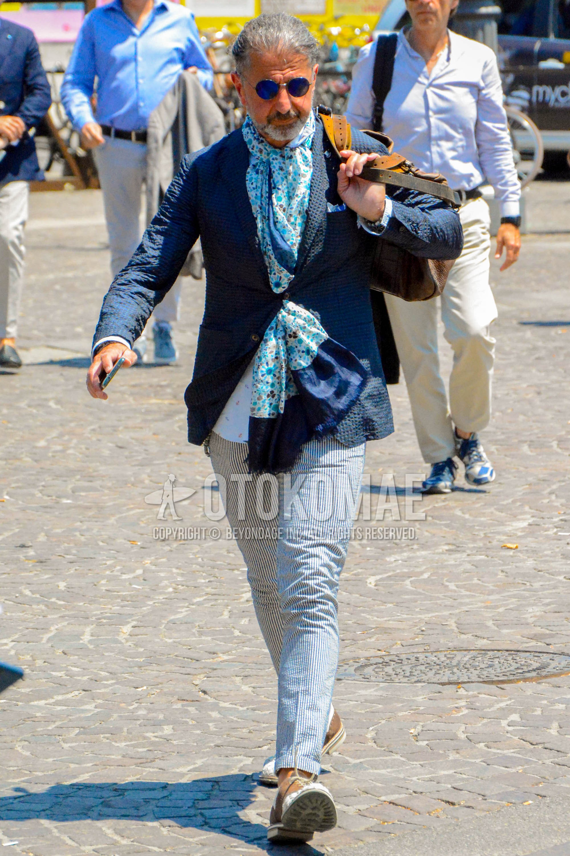Men's spring summer autumn outfit with plain sunglasses, light blue scarf scarf, navy plain tailored jacket, white navy graphic slacks.