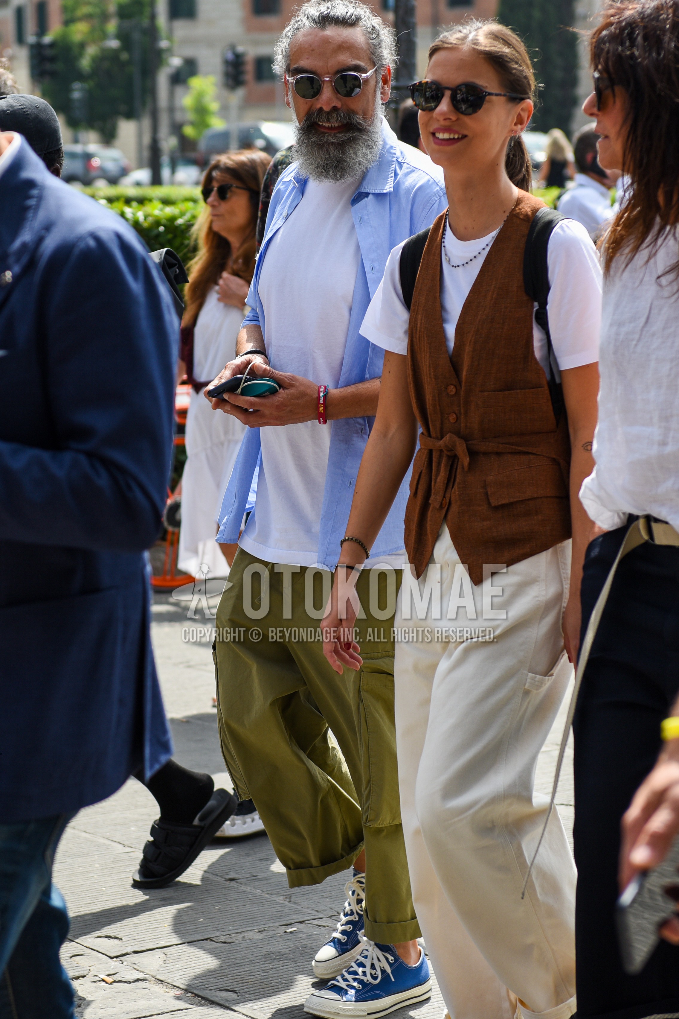 Men's spring summer outfit with clear plain sunglasses, light blue plain shirt, white plain t-shirt, olive green plain cargo pants, blue low-cut sneakers.