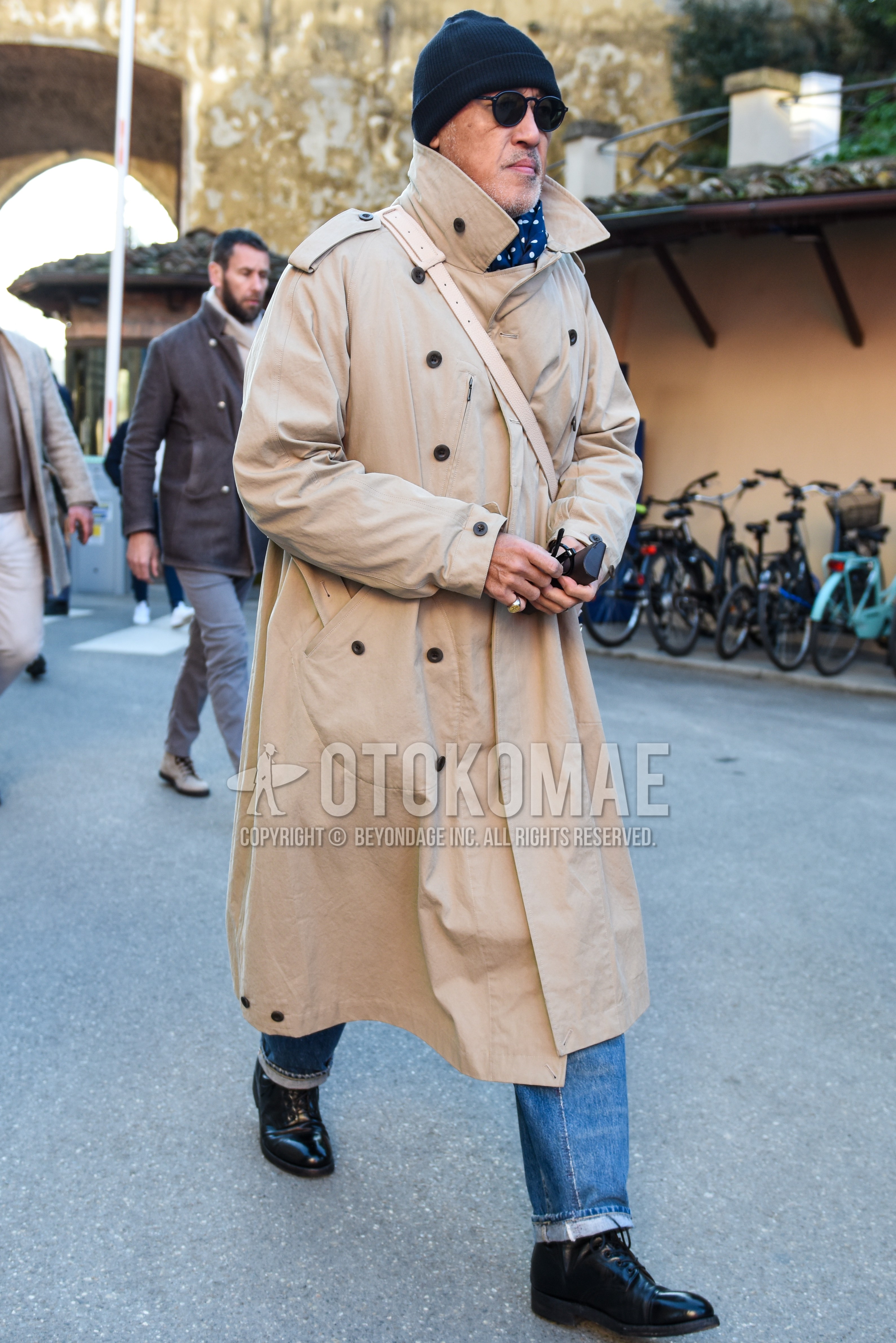 Men's autumn winter outfit with black plain knit cap, black plain sunglasses, navy dots scarf, beige plain trench coat, light blue plain denim/jeans, black  boots.