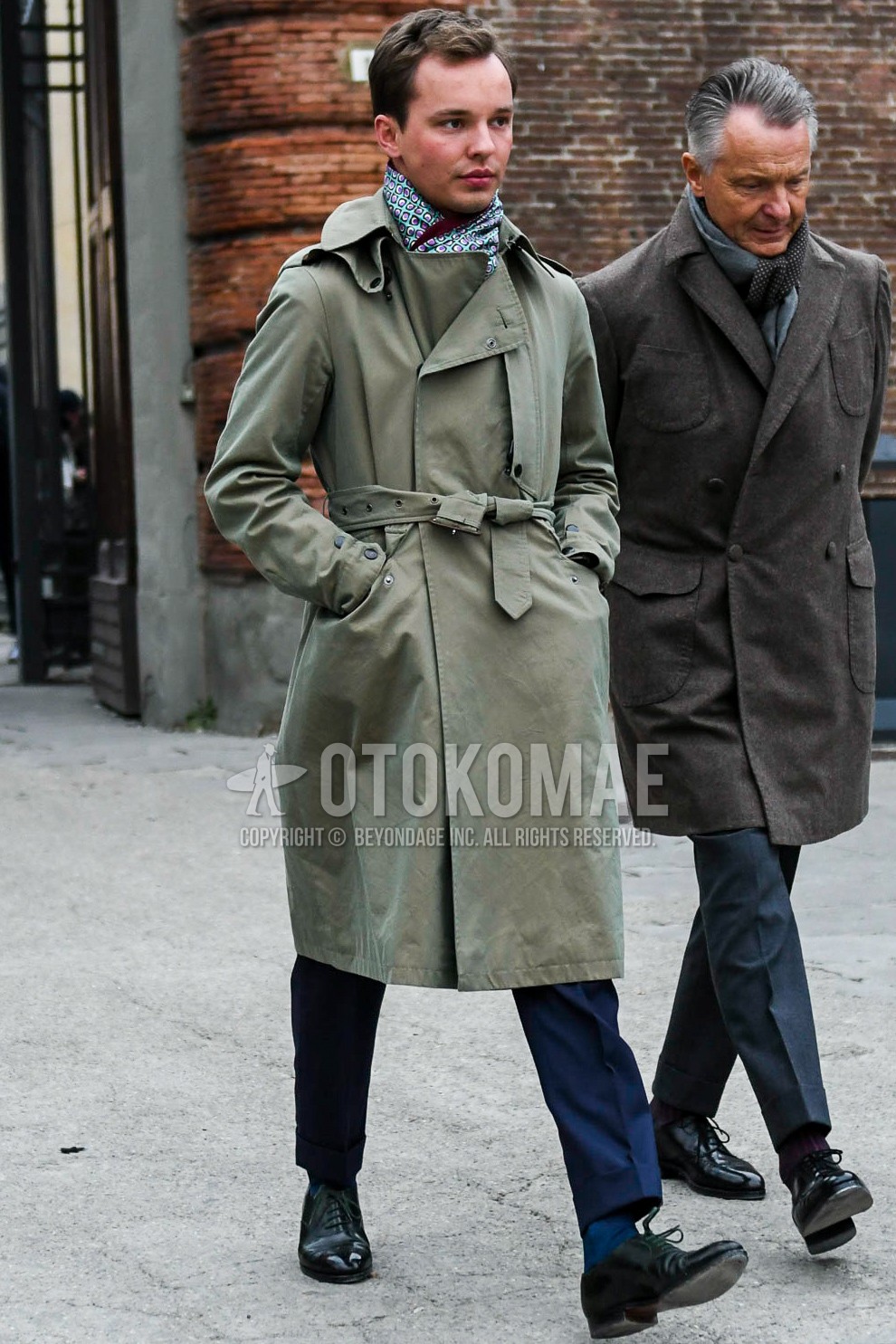 Men's autumn winter outfit with multi-color small crest scarf, olive green plain belted coat, olive green plain trench coat, gray plain slacks, gray plain socks, black plain toe leather shoes.
