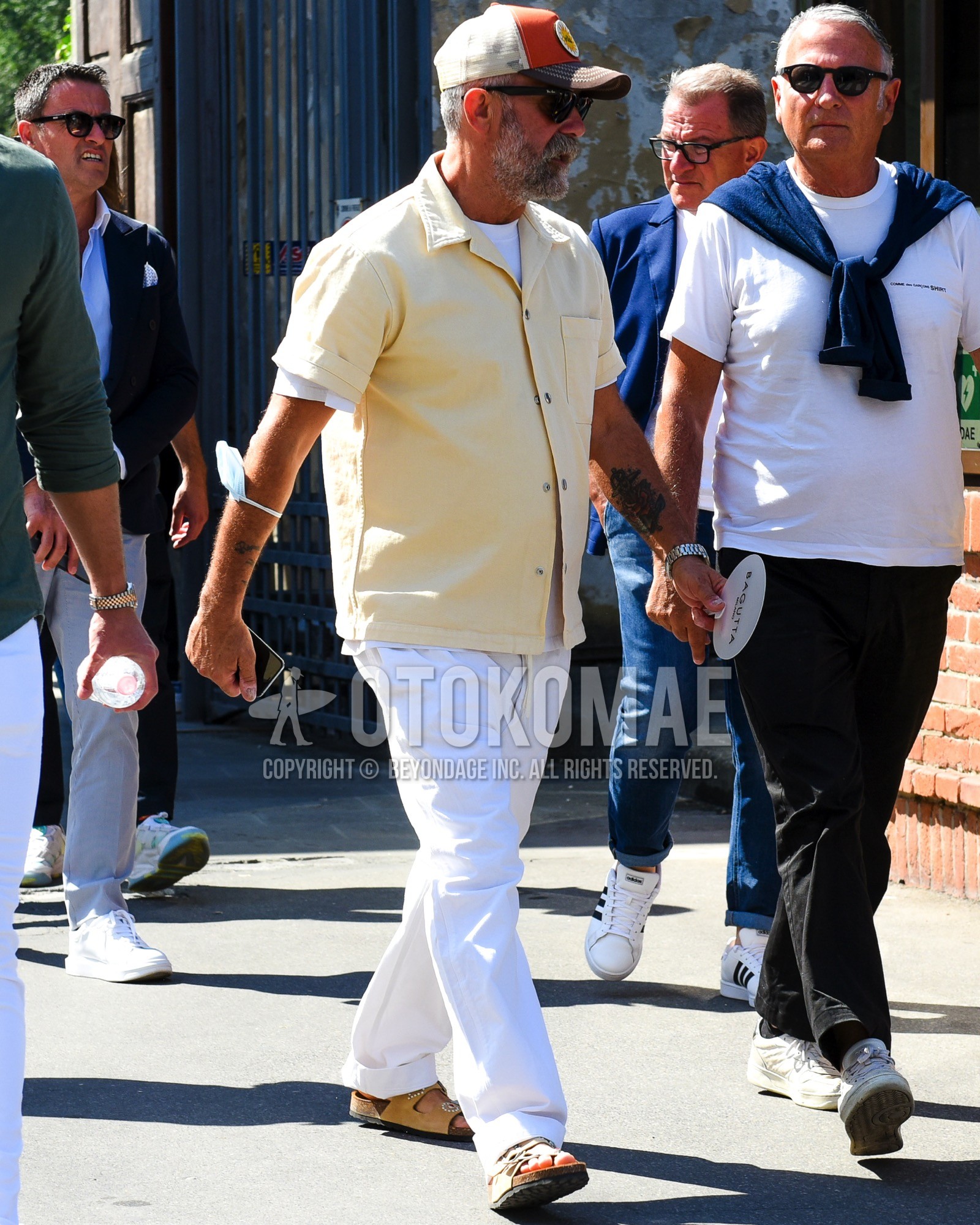 Men's spring summer outfit with orange deca logo baseball cap, plain sunglasses, white plain t-shirt, yellow plain shirt, white plain cotton pants, beige leather sandals.