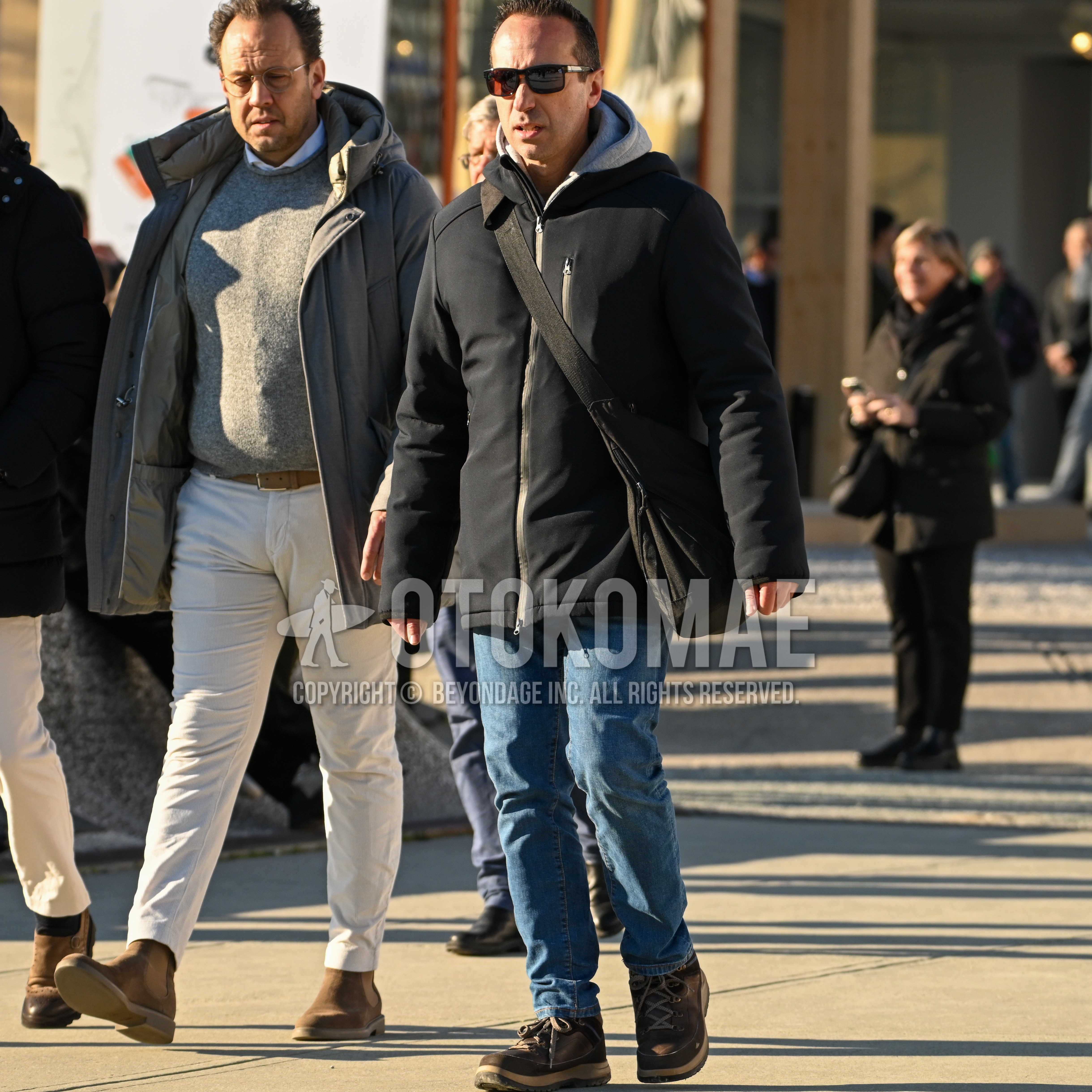 Men's outfit with black plain sunglasses, black plain windbreaker, gray plain hoodie, blue plain denim/jeans, brown high-cut sneakers, black plain shoulder bag.