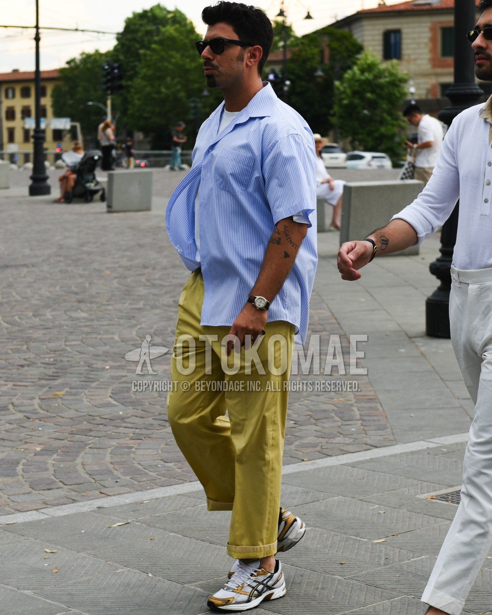 Men's spring summer outfit with black plain sunglasses, blue stripes shirt, white plain t-shirt, yellow plain chinos, white low-cut sneakers.