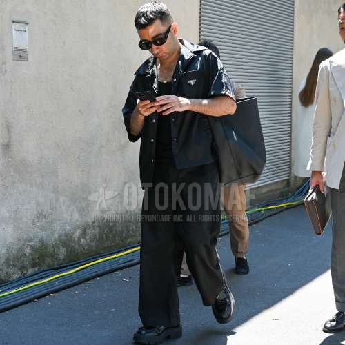 黒無地のサングラス、プラダの黒無地のシャツ、黒無地のタンクトップ、黒無地のスラックス、黒無地のチノパン、プラダの黒ローファーの革靴、レザーの黒無地のトートバッグを合わせた春夏のメンズコーデ・着こなし。