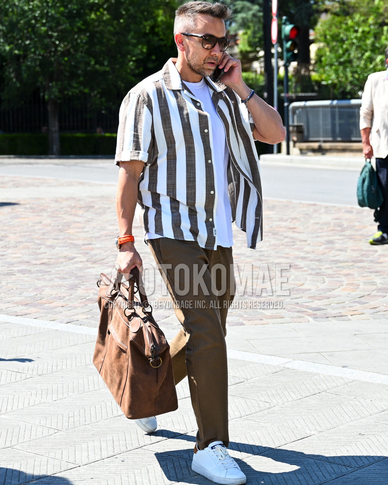 Men's spring summer outfit with brown plain sunglasses, brown white stripes shirt, brown plain chinos, white low-cut sneakers, brown plain briefcase/handbag.