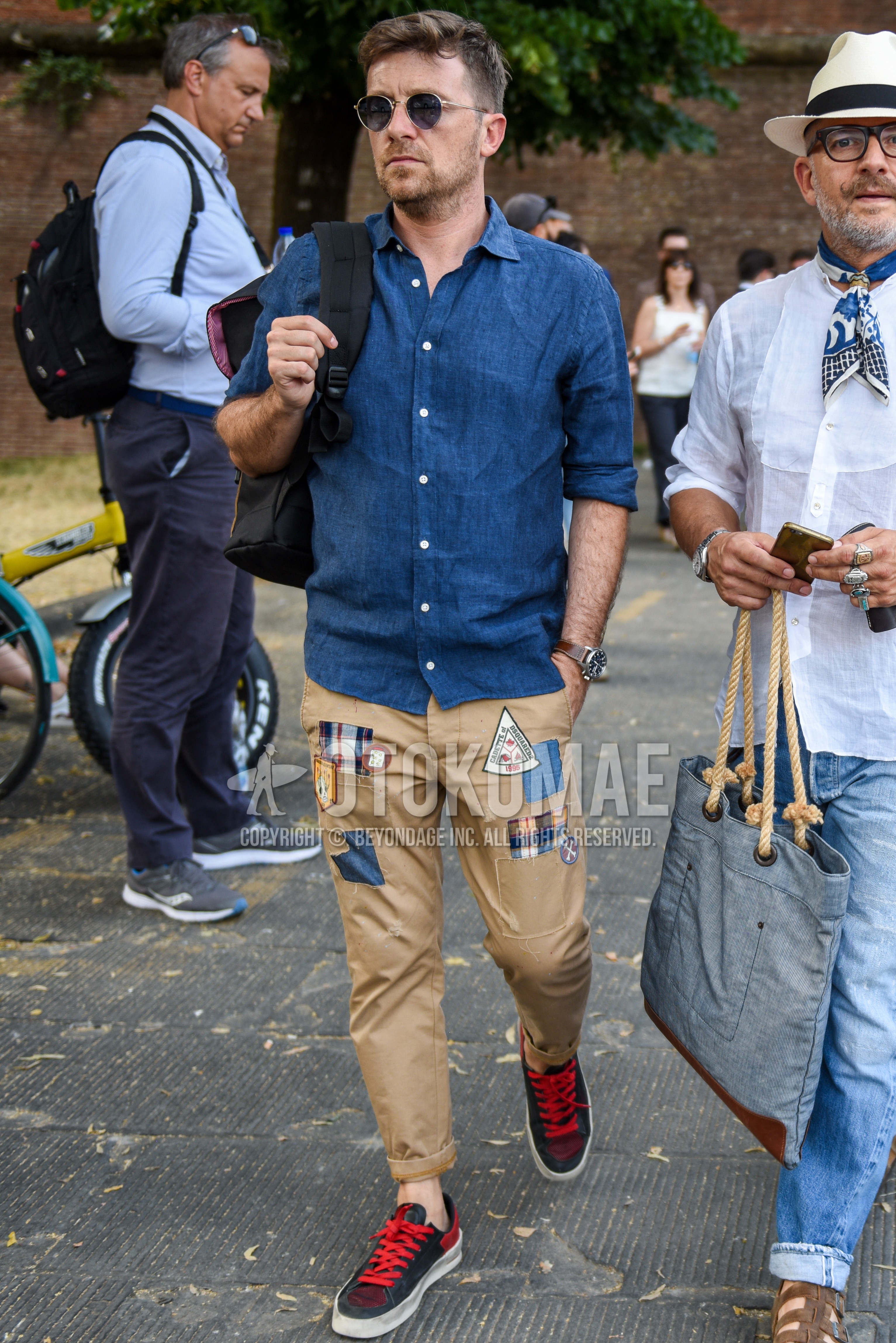 Men's spring summer outfit with black gold plain sunglasses, blue plain shirt, beige plain chinos, black red low-cut sneakers.