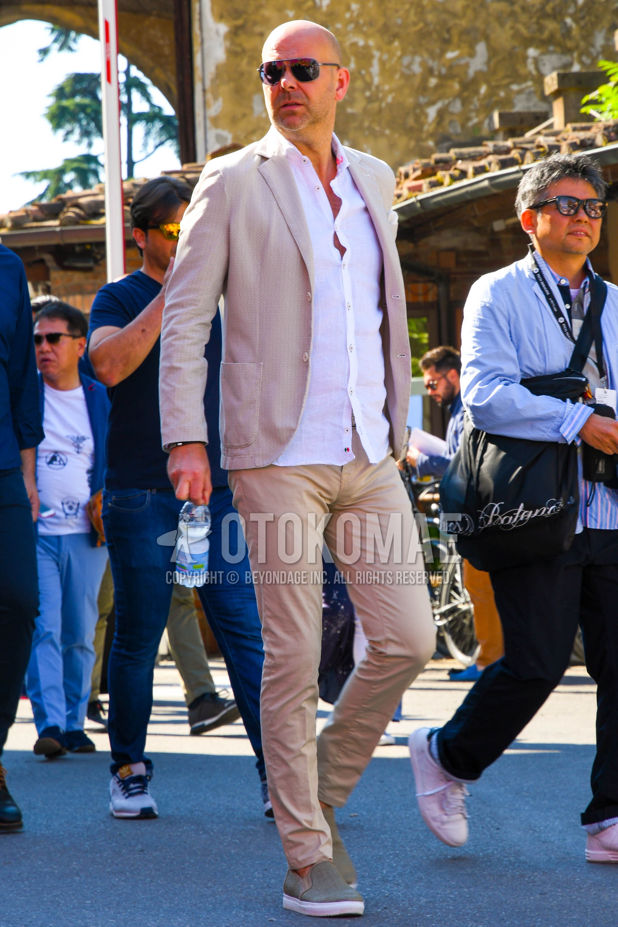 Men's spring summer autumn outfit with silver plain sunglasses, beige plain tailored jacket, white plain shirt, beige plain chinos, beige slip-on sneakers.