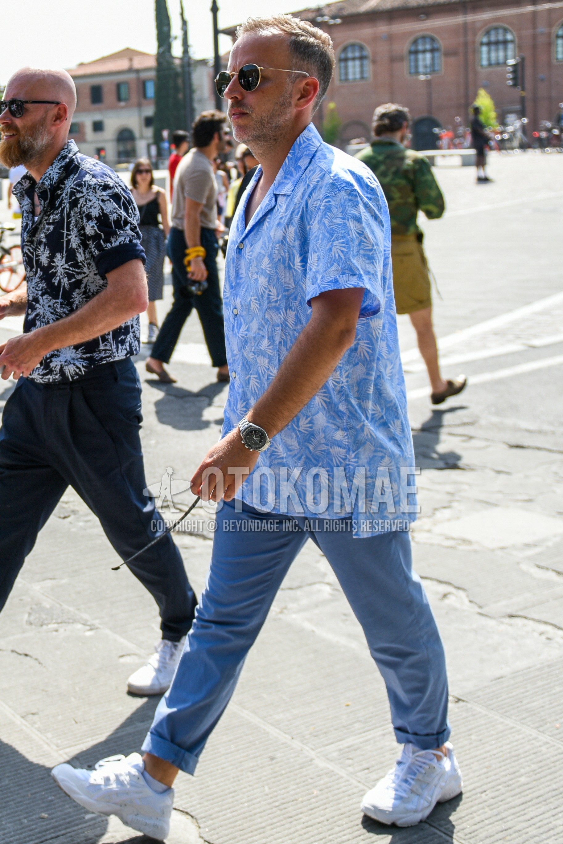 Men's summer outfit with gold plain sunglasses, light blue botanical shirt, light blue plain cotton pants, white low-cut sneakers.