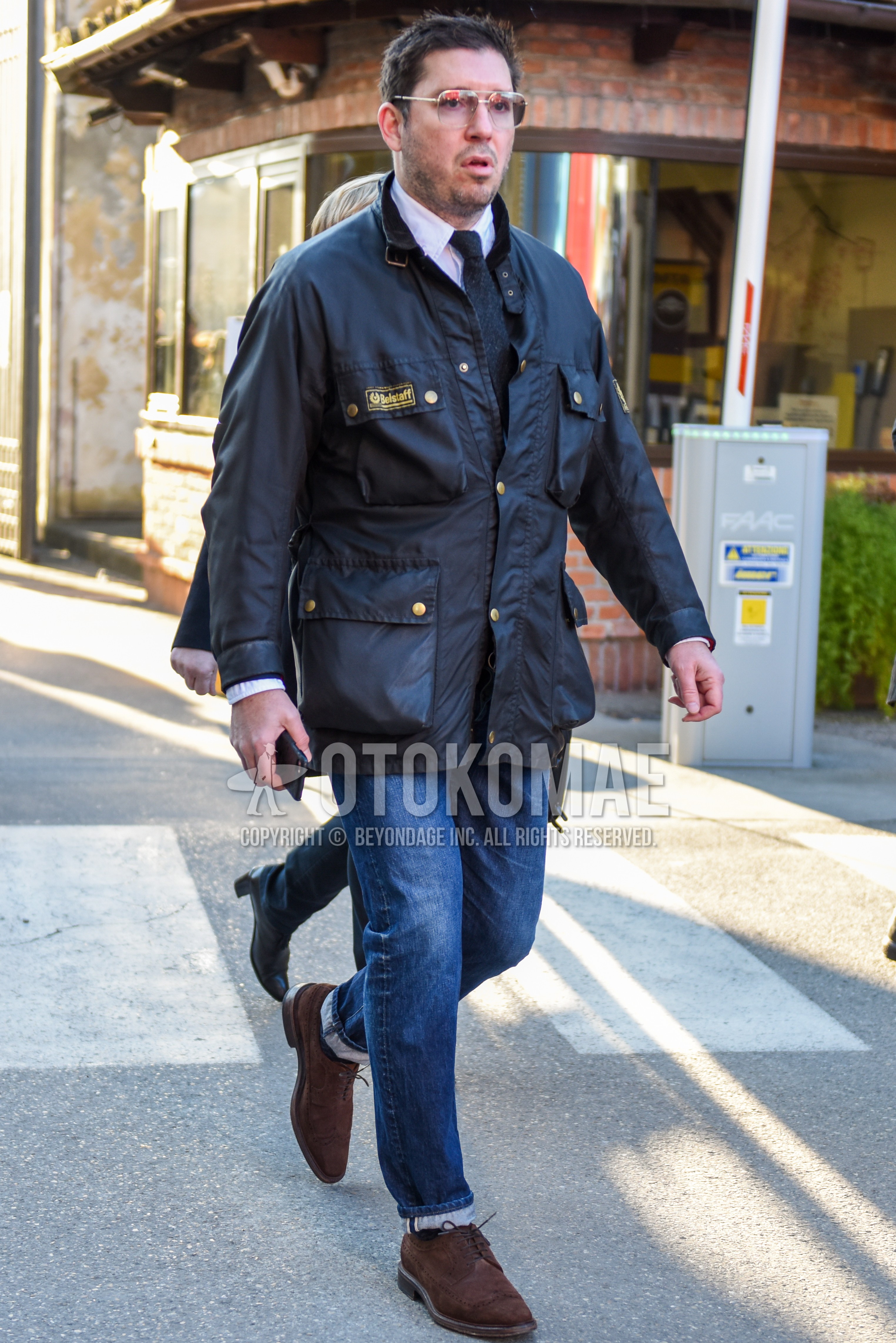 Men's autumn winter outfit with gold plain glasses, black plain riders jacket, white plain shirt, blue plain denim/jeans, brown wing-tip shoes leather shoes, gray plain necktie.