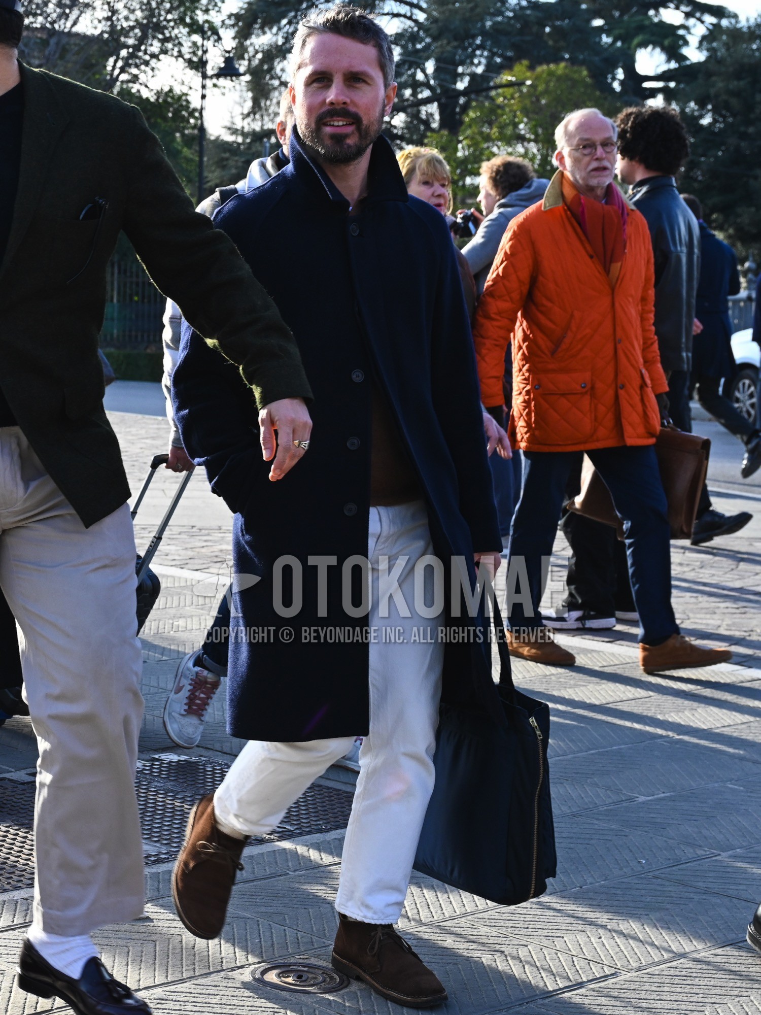Men's autumn winter outfit with navy plain stenkarrer coat, brown plain polo shirt, white plain cotton pants, brown chukka boots, black plain briefcase/handbag.