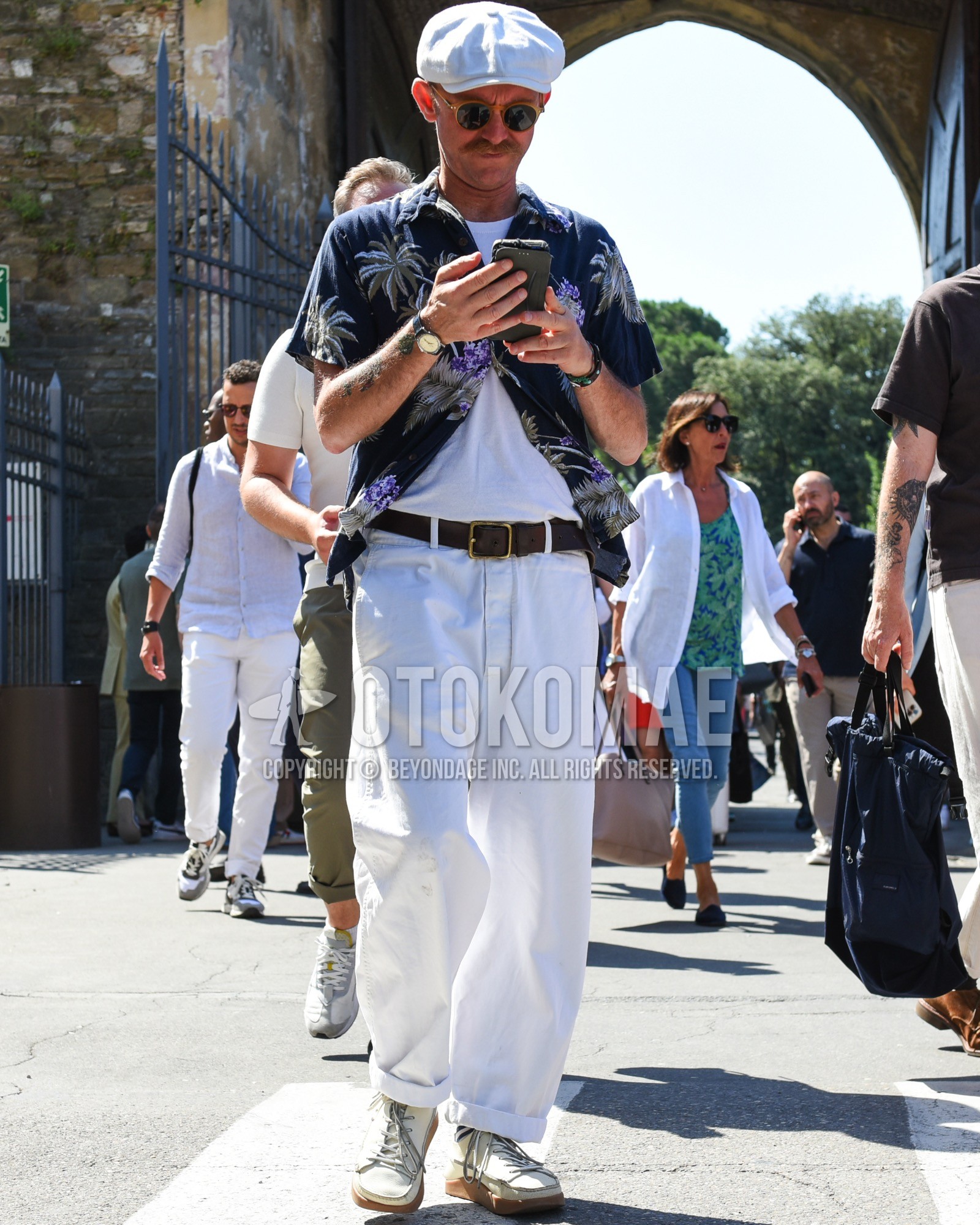 Men's spring summer outfit with white plain hunting cap, brown plain sunglasses, navy botanical shirt, white plain t-shirt, brown plain leather belt, white plain cotton pants, navy white horizontal stripes socks, beige low-cut sneakers.