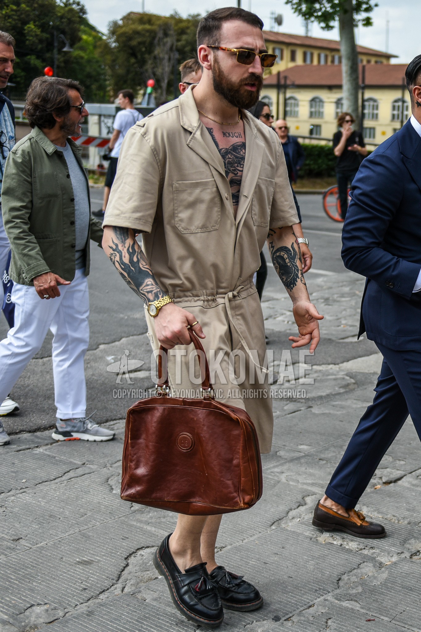 Men's summer outfit with brown tortoiseshell sunglasses, beige plain jumpsuit, black tassel loafers leather shoes, brown plain briefcase/handbag.
