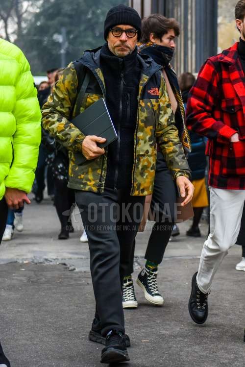 黒無地のニットキャップ、無地のメガネ、ノースフェイスのオリーブグリーン迷彩のマウンテンパーカー、黒無地のフリースジャケット、グレー無地のスラックス、黒ローカットのスニーカーを合わせた冬のメンズコーデ・着こなし。