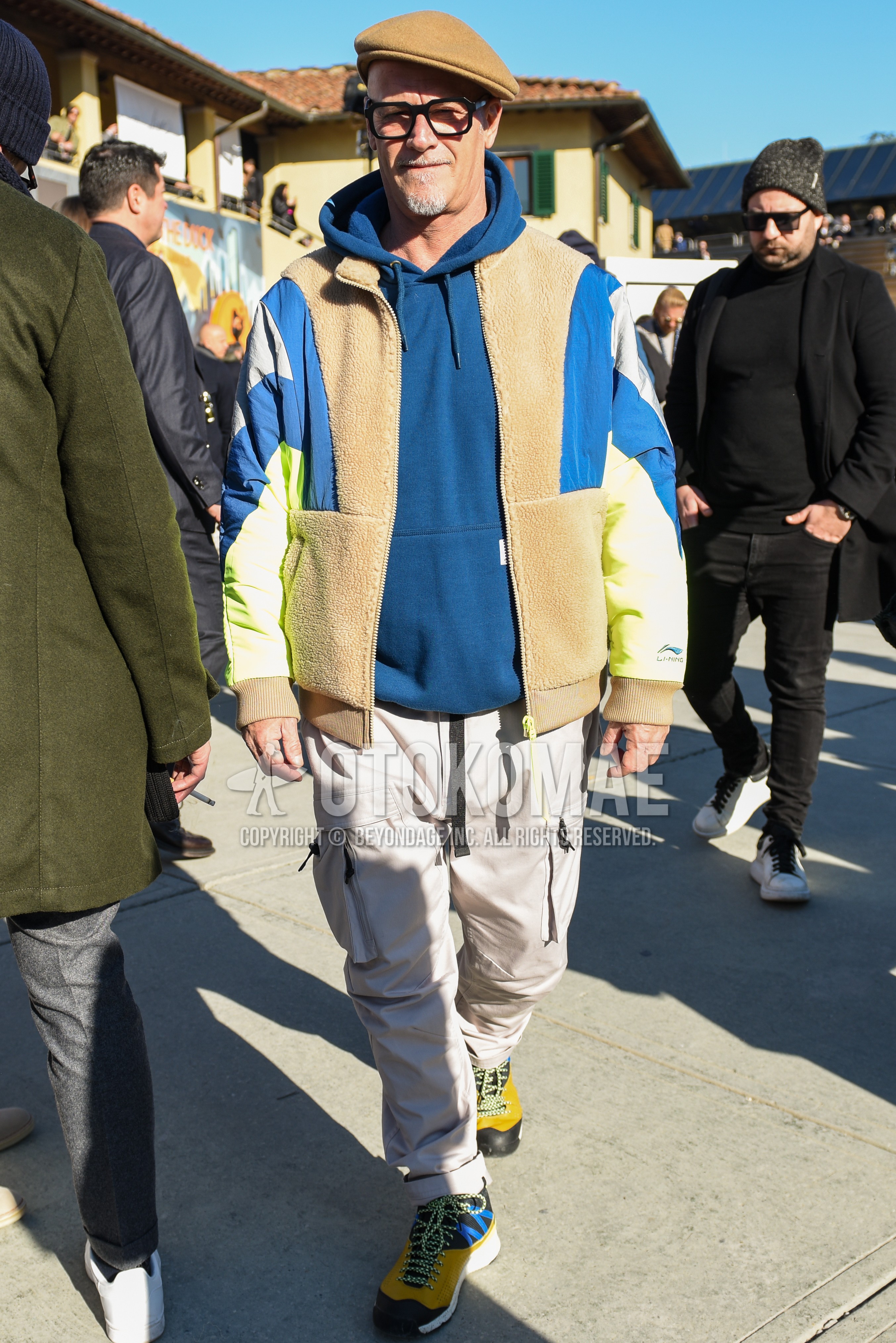 Men's autumn winter outfit with beige plain hunting cap, black plain glasses, multi-color plain fleece jacket, navy plain hoodie, beige plain cargo pants, yellow black high-cut sneakers.