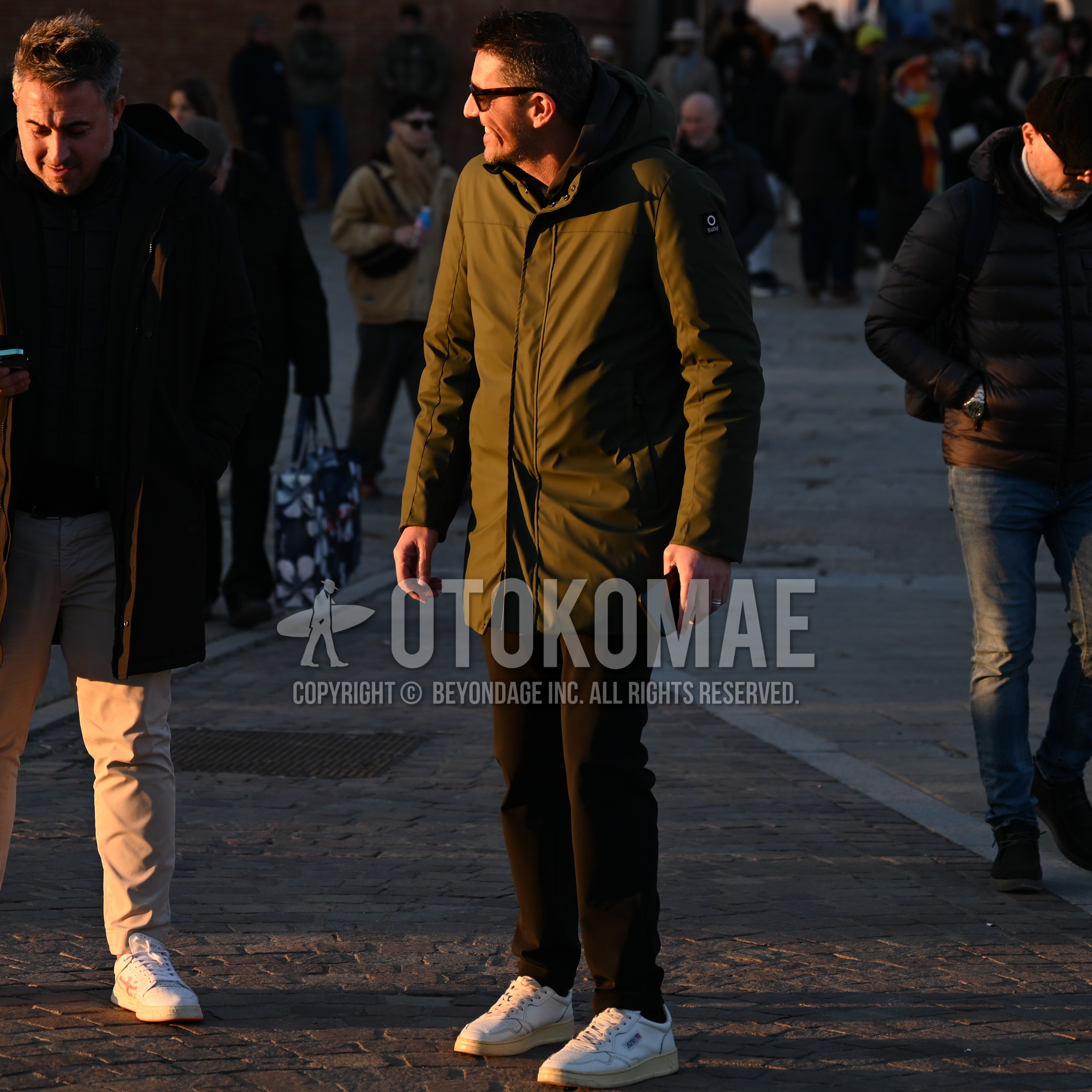 Men's autumn winter outfit with black plain sunglasses, olive green plain hooded coat, black plain chinos, white low-cut sneakers.
