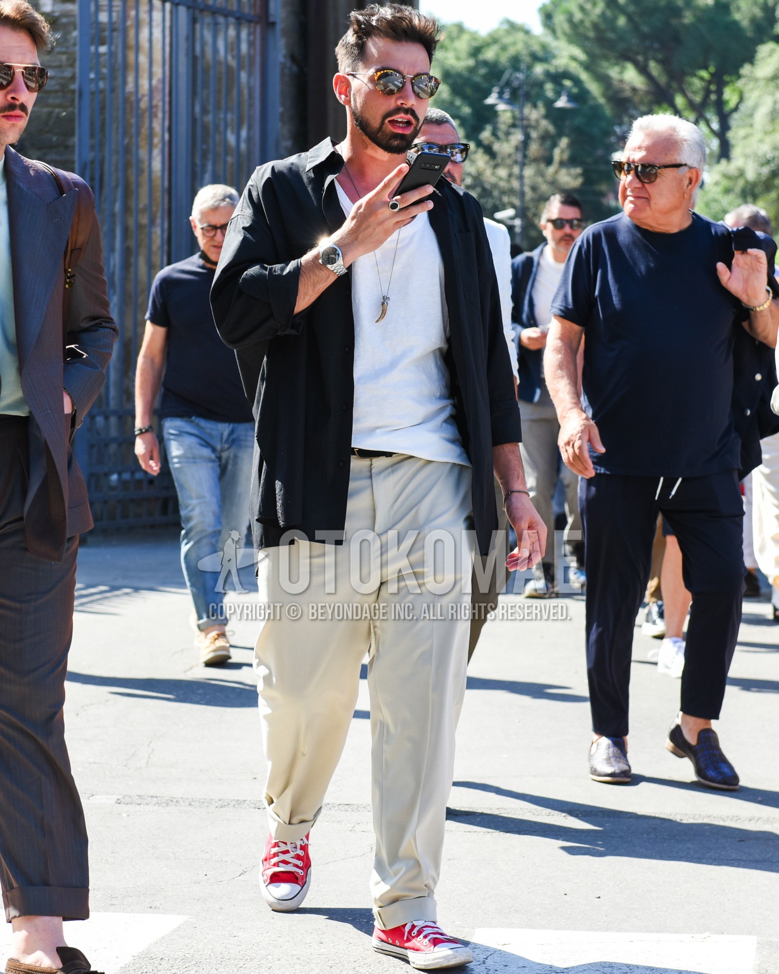 Men's spring summer outfit with brown tortoiseshell sunglasses, black plain shirt, white plain t-shirt, black plain leather belt, beige plain slacks, red low-cut sneakers.