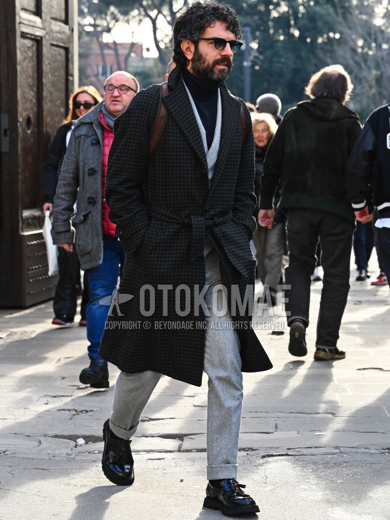 Men's autumn winter outfit with black plain sunglasses, dark gray check belted coat, navy plain turtleneck knit, gray plain tailored jacket, gray plain slacks, black plain socks, black tassel loafers leather shoes, brown plain backpack.
