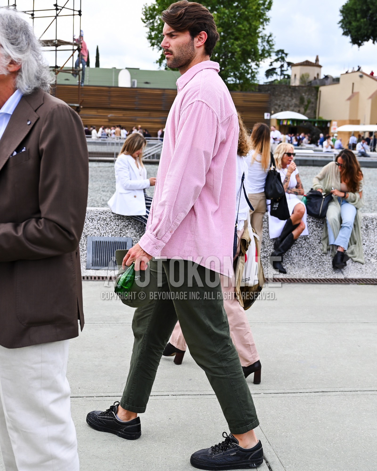Men's spring summer autumn outfit with pink plain shirt, olive green plain chinos, black low-cut sneakers.