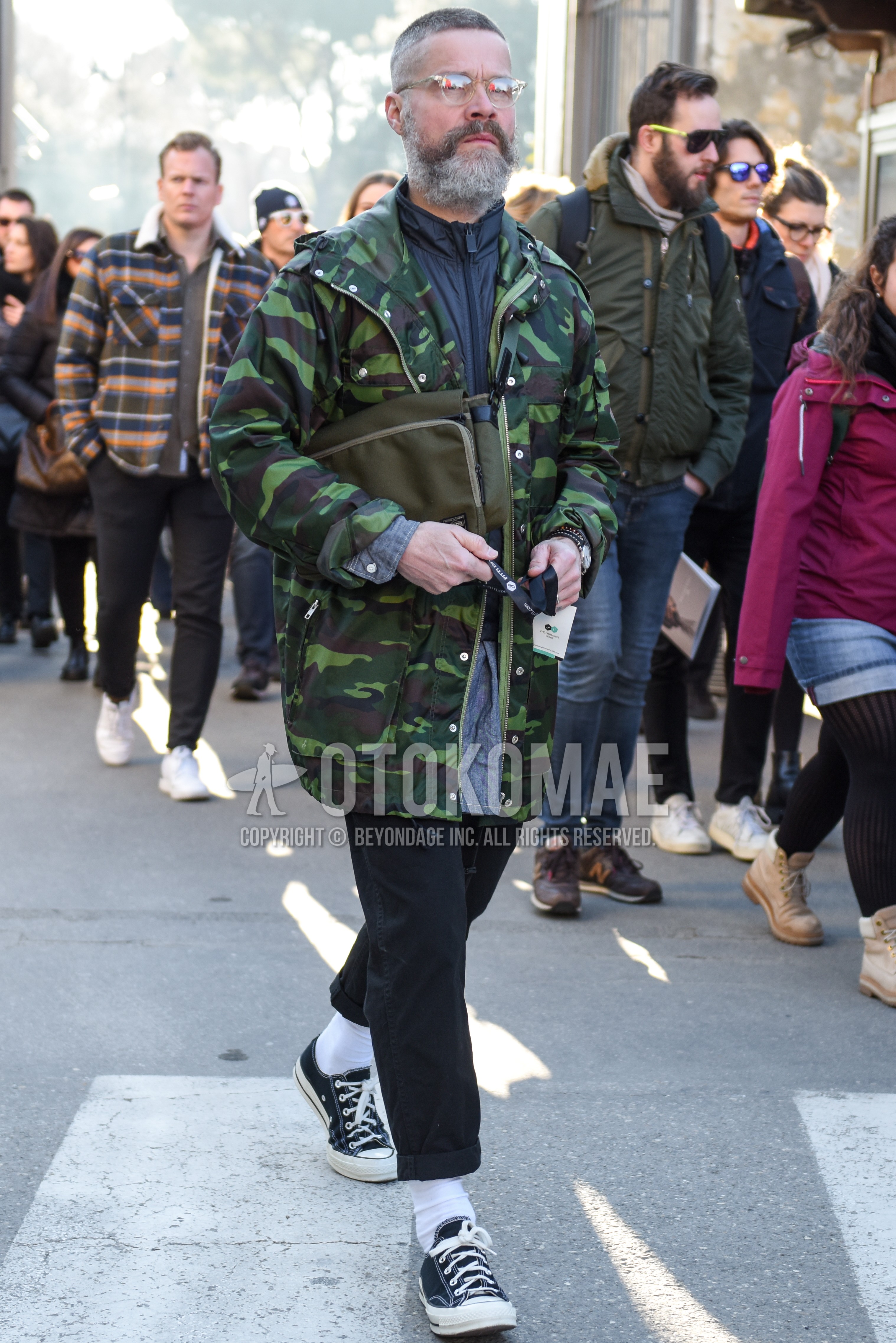 Men's autumn winter outfit with clear plain glasses, olive green camouflage hooded coat, gray plain windbreaker, blue plain denim shirt/chambray shirt, black plain denim/jeans, white plain socks, black low-cut sneakers, olive green plain shoulder bag.