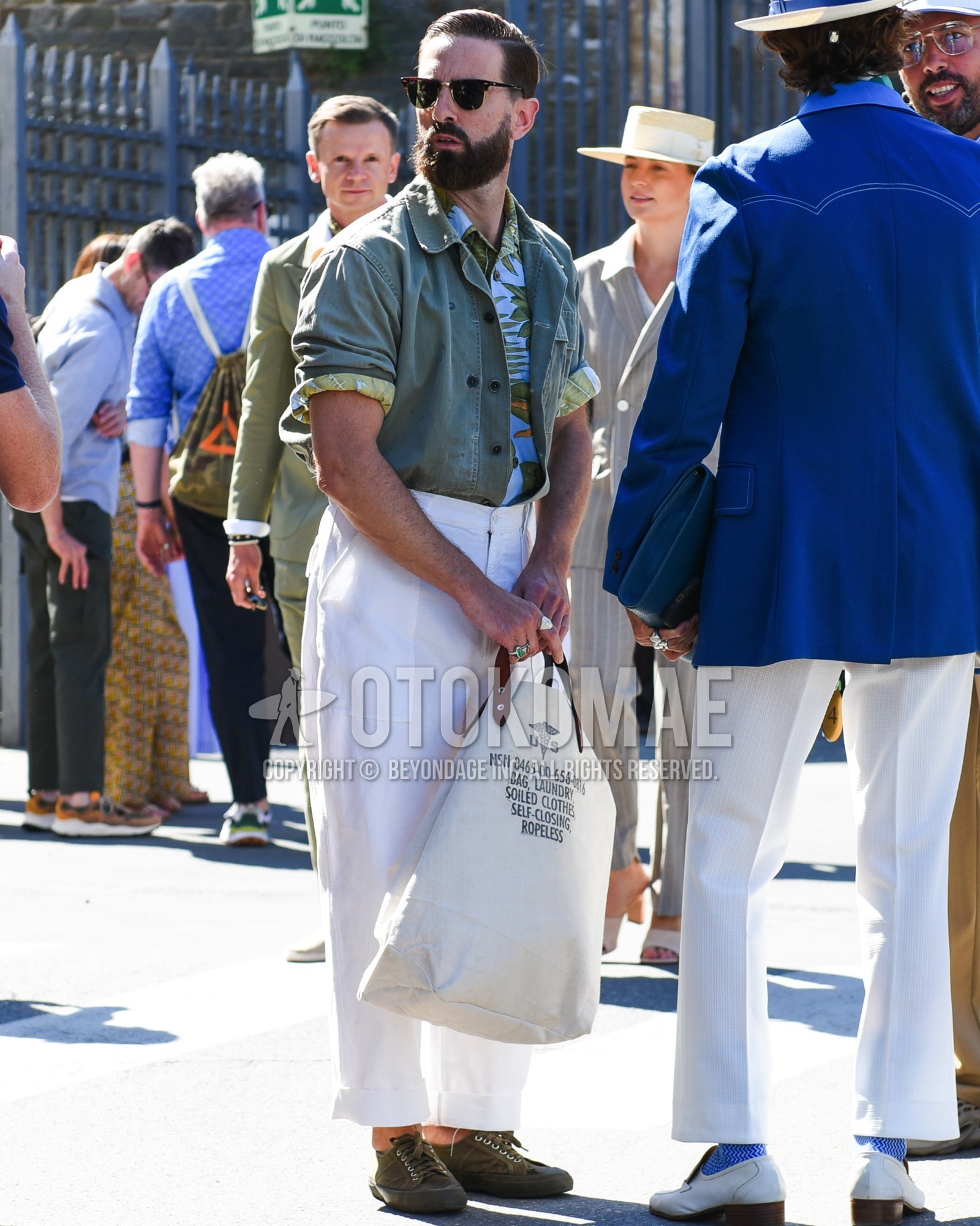 Men's spring summer outfit with brown plain sunglasses, olive green plain shirt jacket, white olive green botanical shirt, white plain baker pants, olive green low-cut sneakers, white deca logo tote bag.