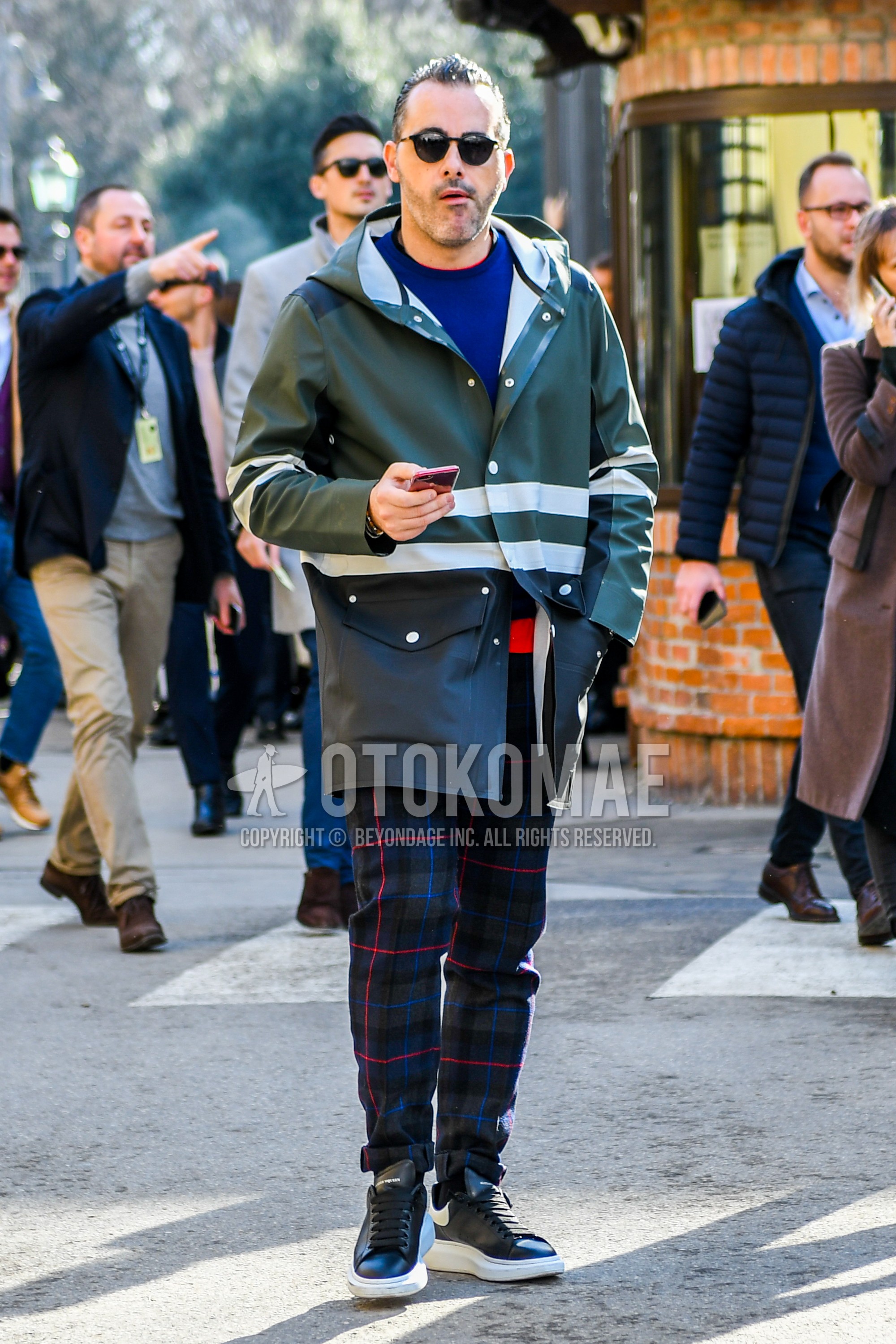Men's autumn winter outfit with plain sunglasses, olive green black plain hooded coat, blue plain sweater, multi-color check slacks, black low-cut sneakers.