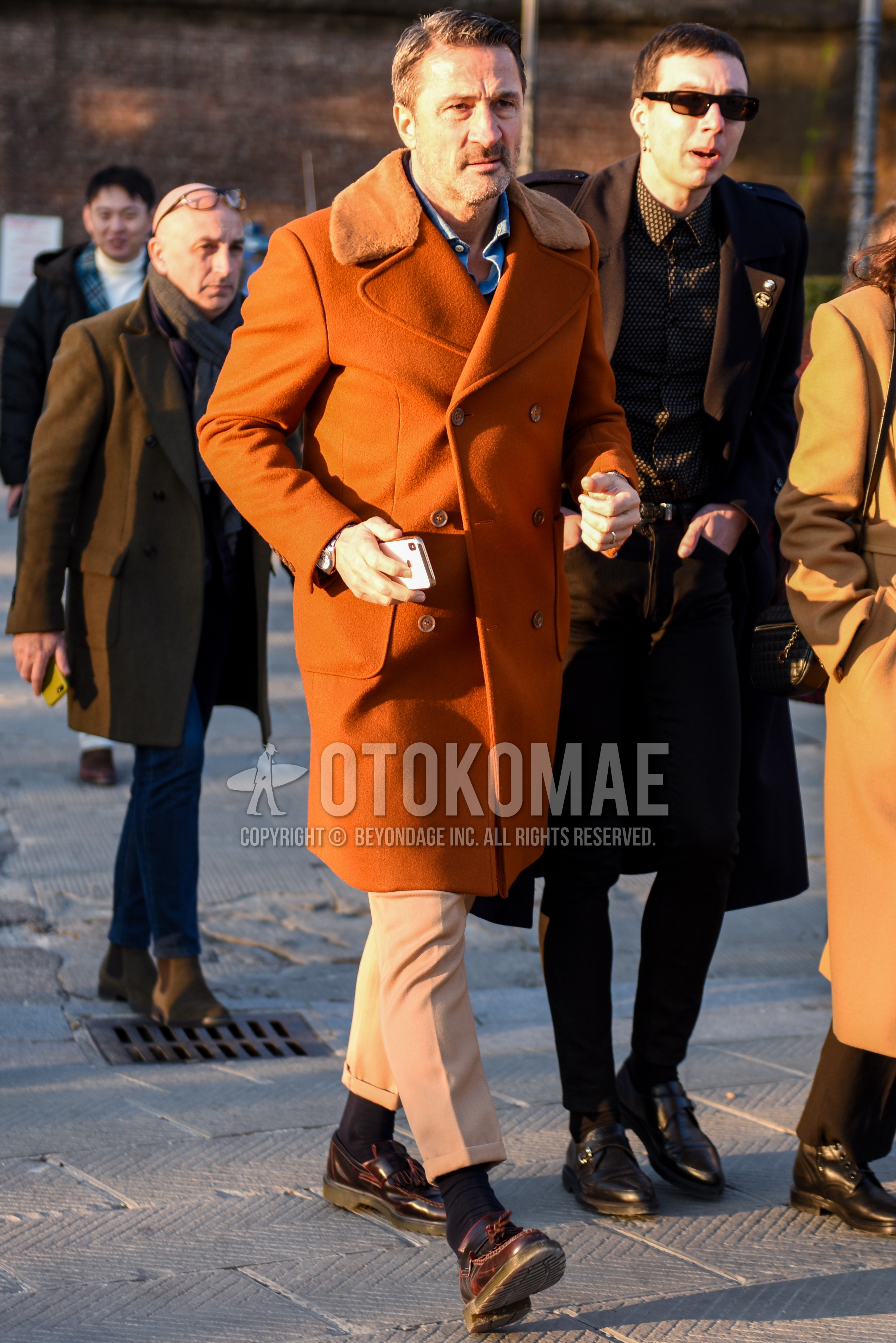 Men's autumn winter outfit with orange plain ulster coat, light blue plain shirt, beige plain slacks, black plain socks, brown tassel loafers leather shoes.
