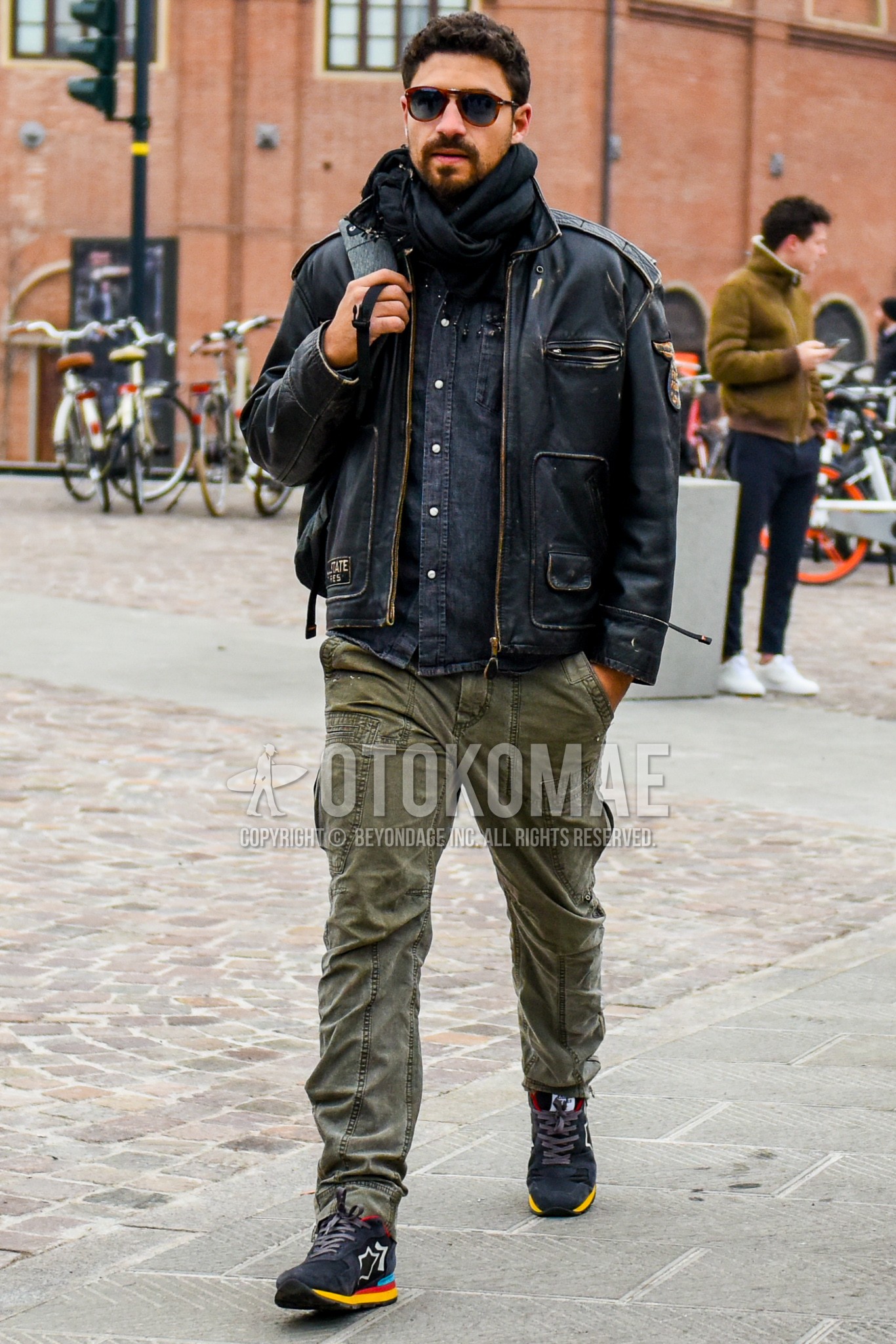 Men's winter outfit with plain sunglasses, black plain scarf, black plain leather jacket, black plain denim shirt/chambray shirt, olive green plain cargo pants, dark gray low-cut sneakers.