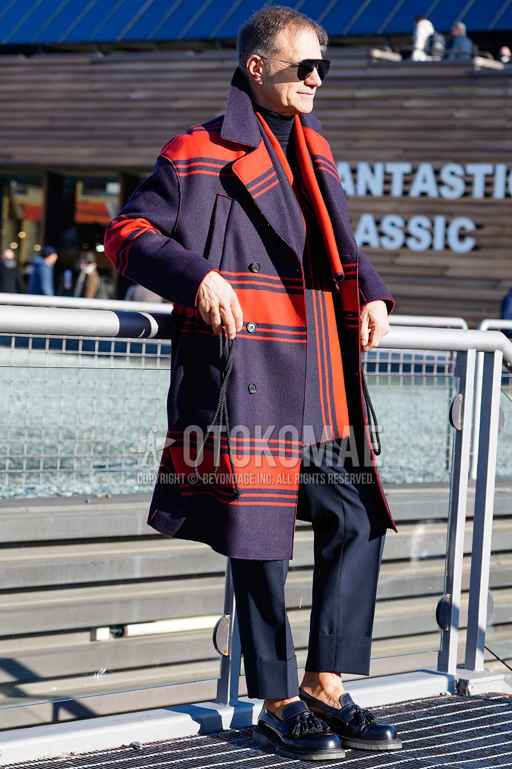 Men's spring winter outfit with black plain sunglasses, red navy horizontal stripes scarf, red navy horizontal stripes ulster coat, navy black plain turtleneck knit, navy plain slacks, black tassel loafers leather shoes.