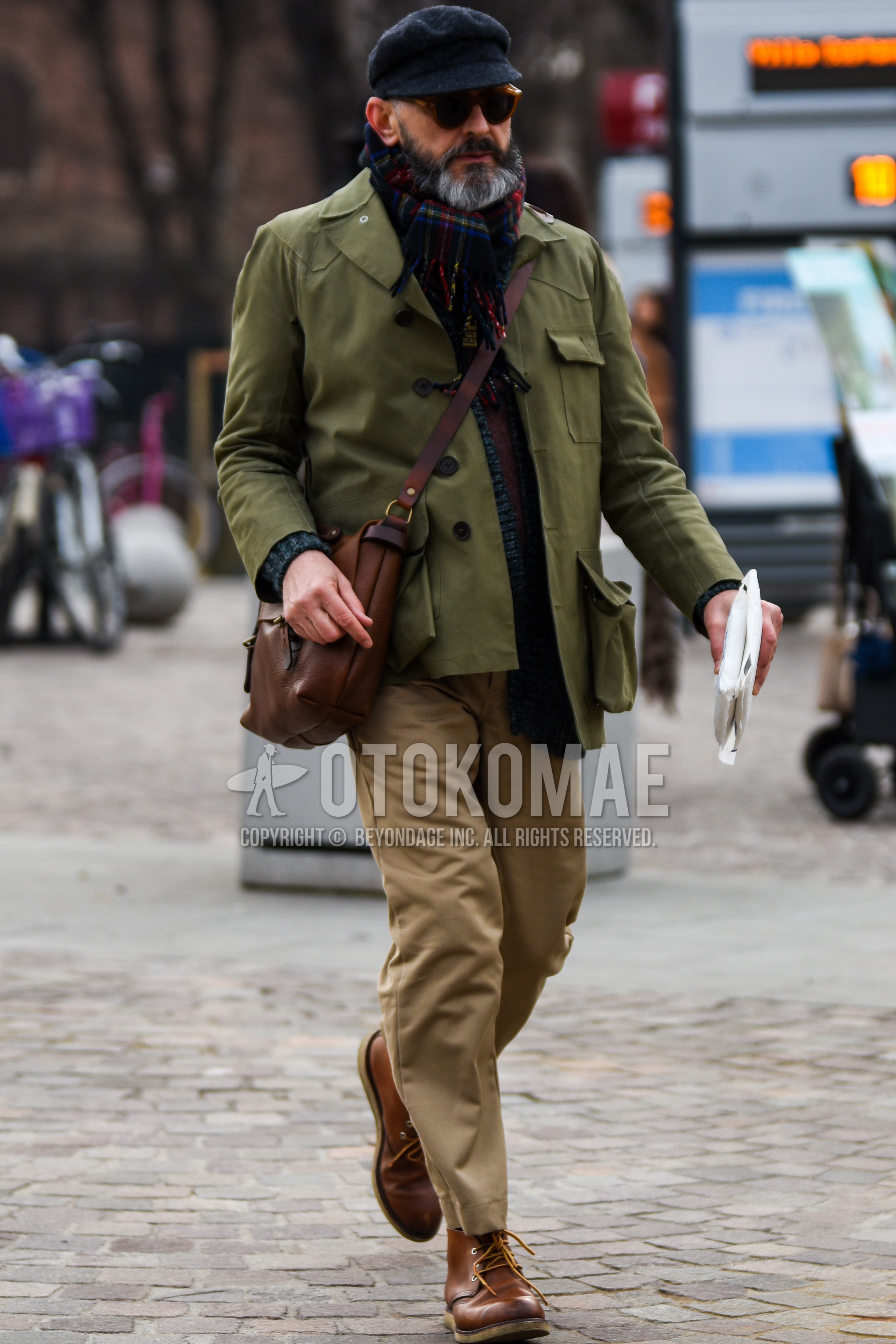 Men's autumn winter outfit with black plain hunting cap, brown tortoiseshell sunglasses, multi-color check scarf, olive green plain shirt jacket, beige plain chinos, brown  boots.