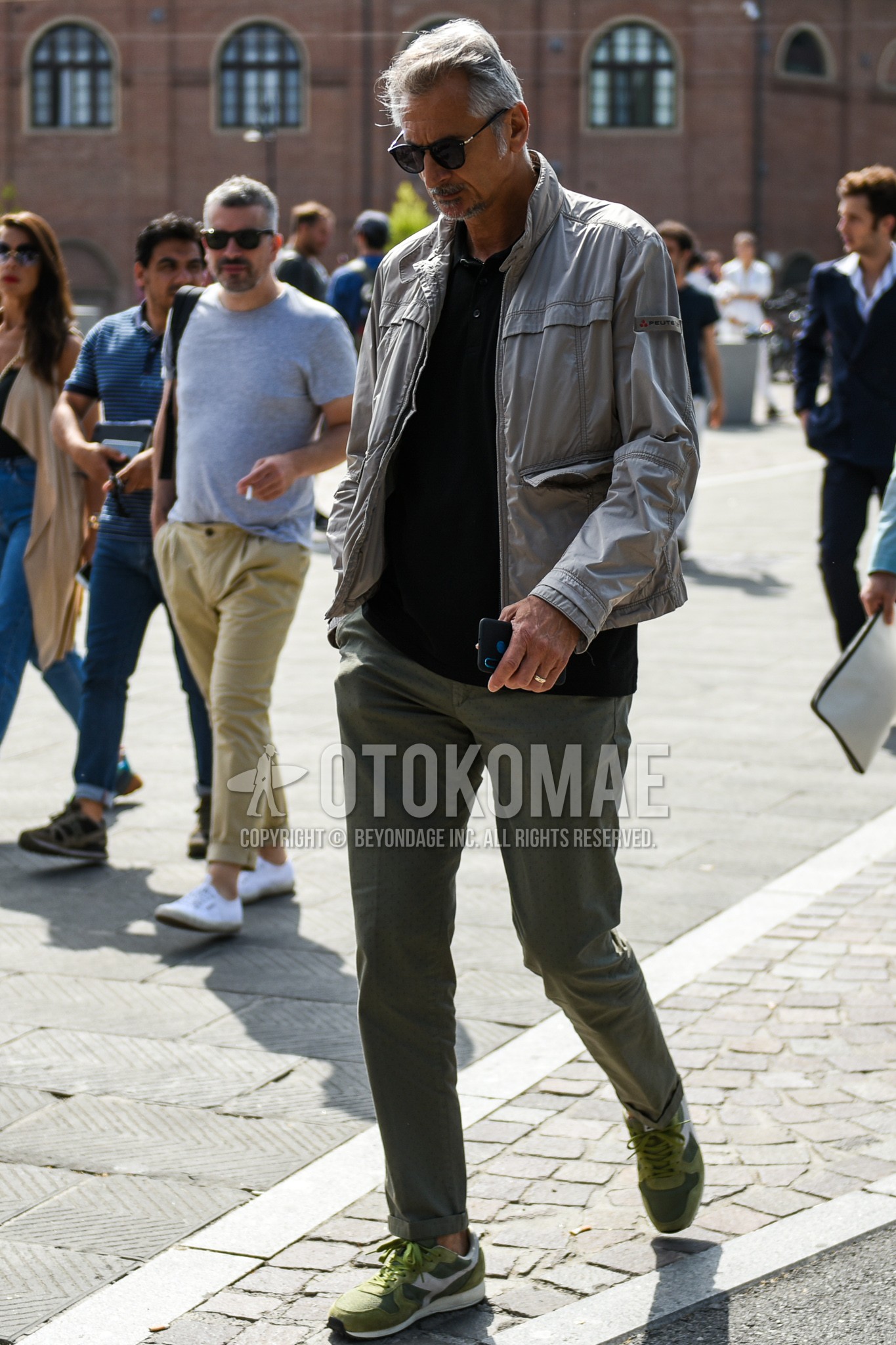 Men's spring autumn outfit with black plain sunglasses, gray plain windbreaker, black plain polo shirt, olive green plain chinos, olive green low-cut sneakers.