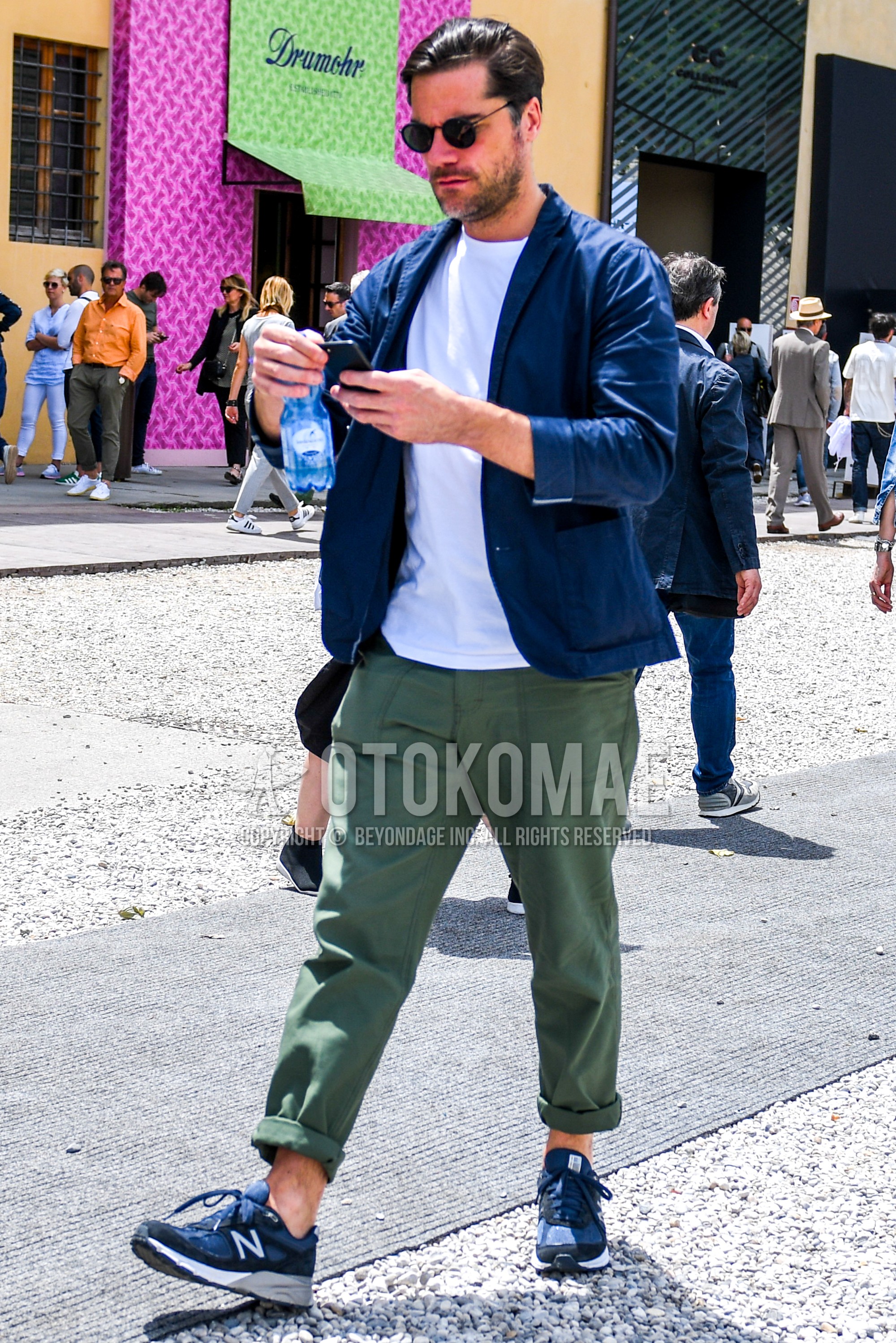 Men's spring summer autumn outfit with plain sunglasses, blue plain tailored jacket, white plain t-shirt, green plain chinos, navy low-cut sneakers.