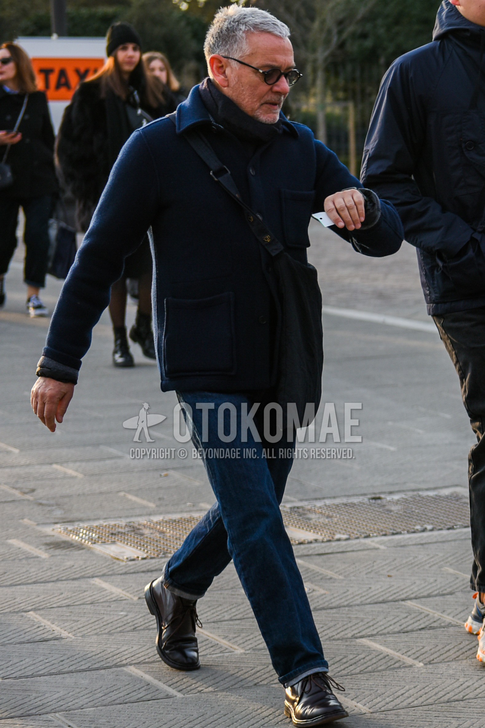 Men's autumn winter outfit with black plain sunglasses, gray plain scarf, navy plain shirt jacket, blue plain denim/jeans, brown chukka boots, black plain shoulder bag.