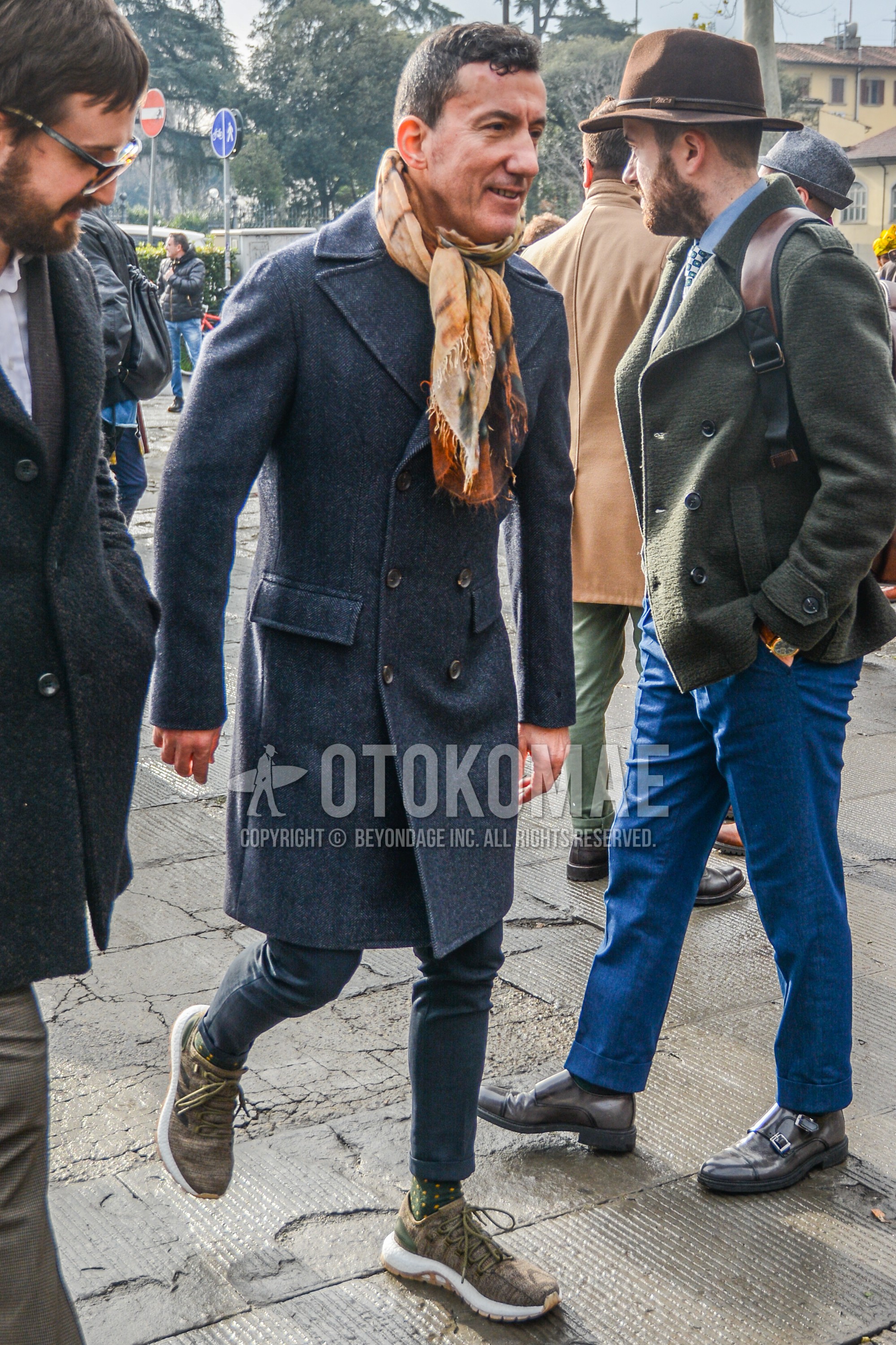 Men's winter outfit with beige scarf scarf, dark gray plain ulster coat, dark gray plain slacks, olive green dots socks, brown low-cut sneakers.
