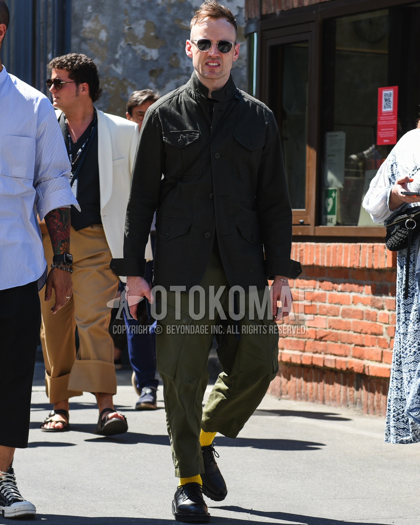 Men's spring summer outfit with clear plain sunglasses, olive green plain field jacket/hunting jacket, olive green plain cargo pants, yellow plain socks, black plain toe leather shoes.