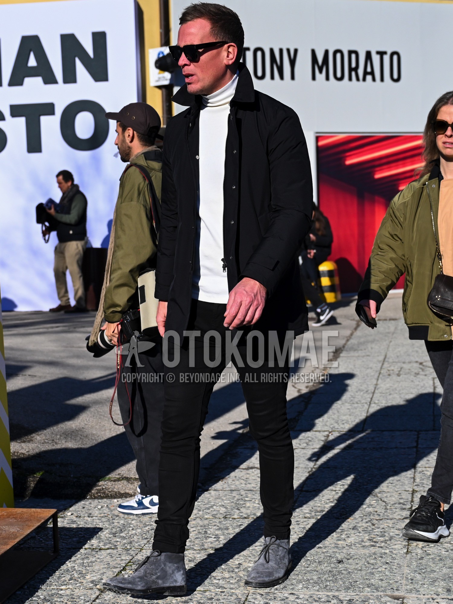 Men's autumn winter outfit with black plain sunglasses, black plain coach jacket, black plain outerwear, white plain turtleneck knit, black plain skinny pants, gray chukka boots.