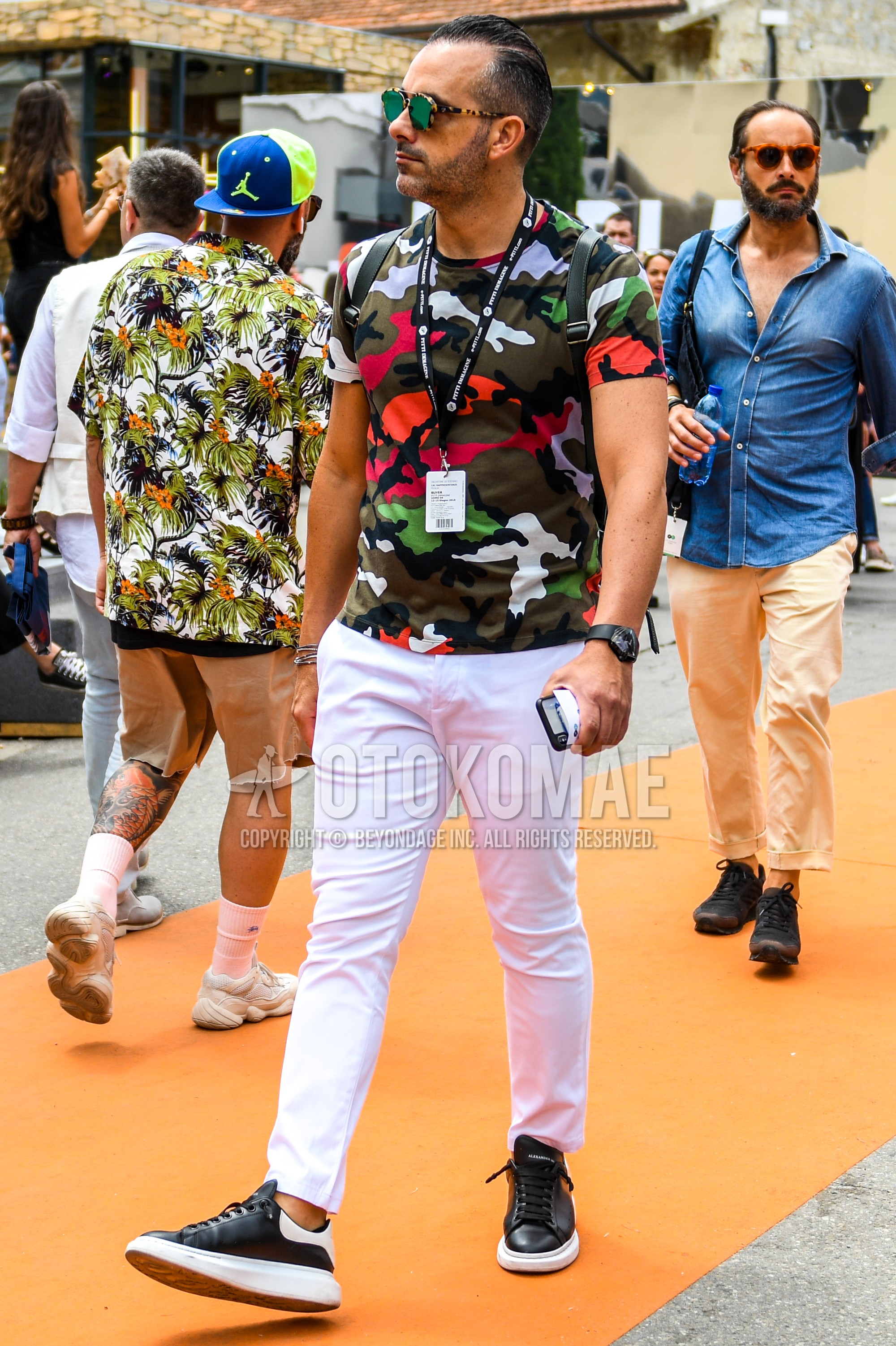 Men's summer outfit with beige brown green tortoiseshell sunglasses, white orange red black camouflage t-shirt, white plain chinos, white plain ankle pants, black white low-cut sneakers, black plain backpack.