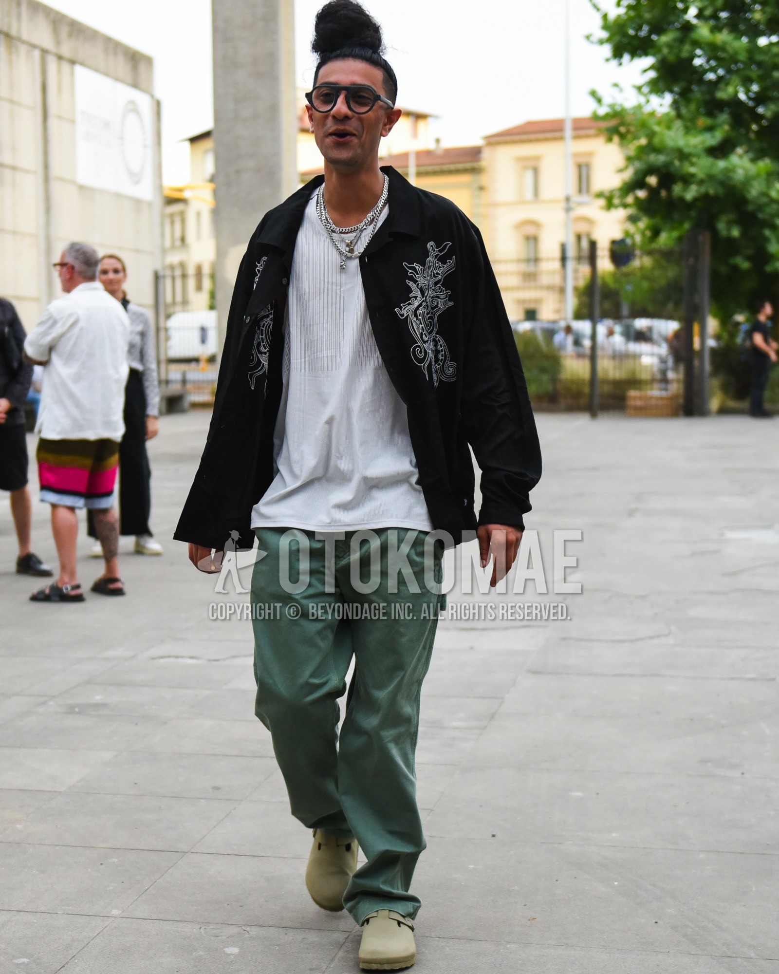 Men's spring summer outfit with black plain glasses, black outerwear coach jacket, white plain t-shirt, olive green plain chinos, beige leather sandals.