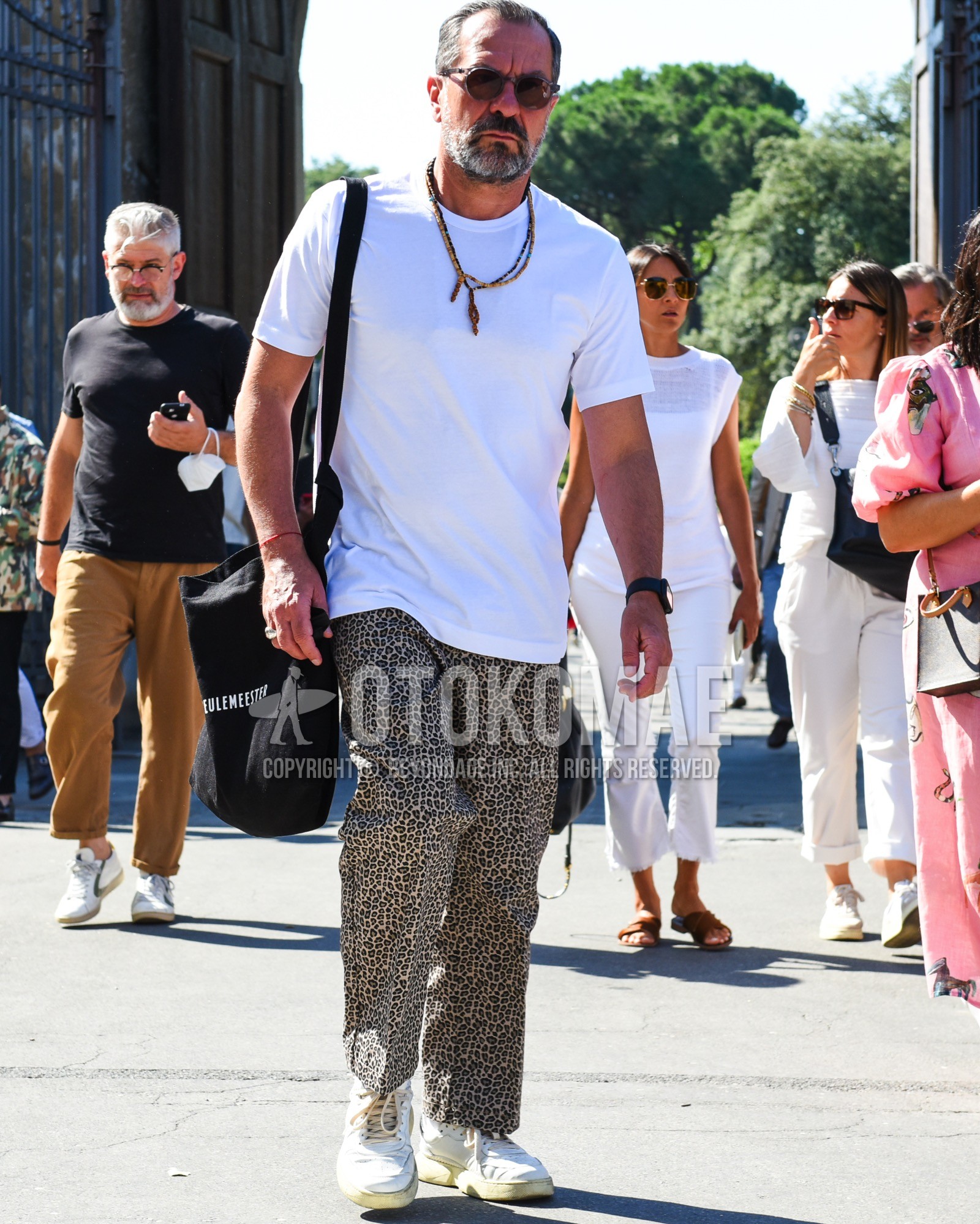 Men's summer outfit with gray tortoiseshell sunglasses, white plain t-shirt, brown leopard slacks, white low-cut sneakers, black deca logo tote bag.